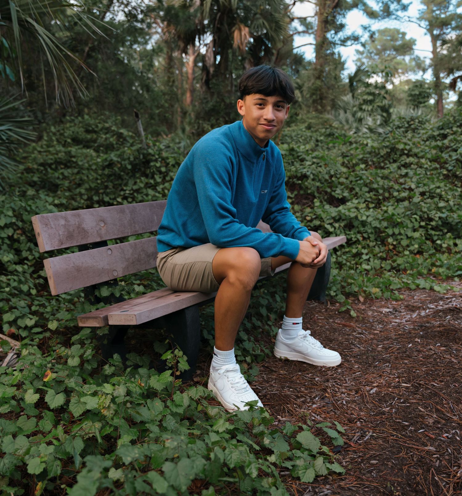 Person in blue jacket sitting on wooden bench surrounded by forest greenery.