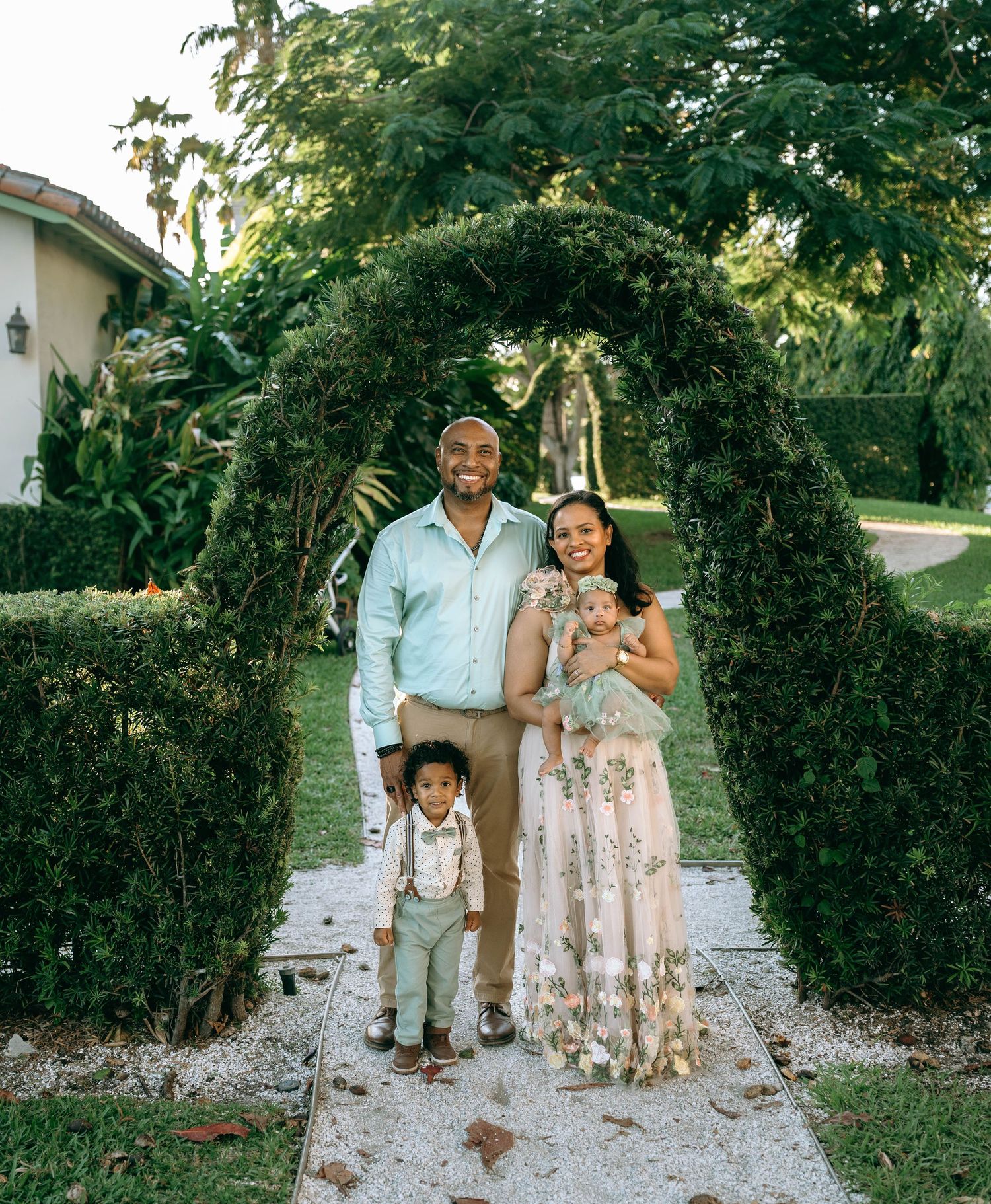 Family of four posing under a lush green archway in a garden, parents smiling with two young children at Bradley Park in Palm Beach, FL.