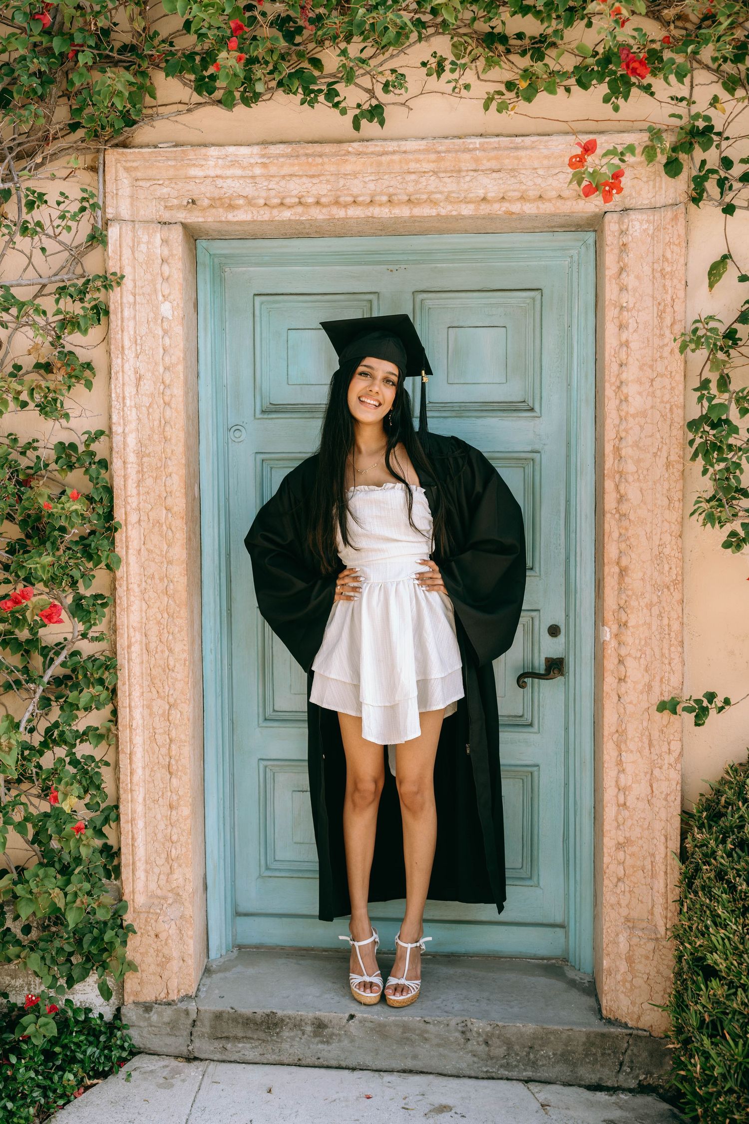Smiling graduate in black cap and gown over white dress standing at a blue door with floral vines on Worth Avenue Palm beach.