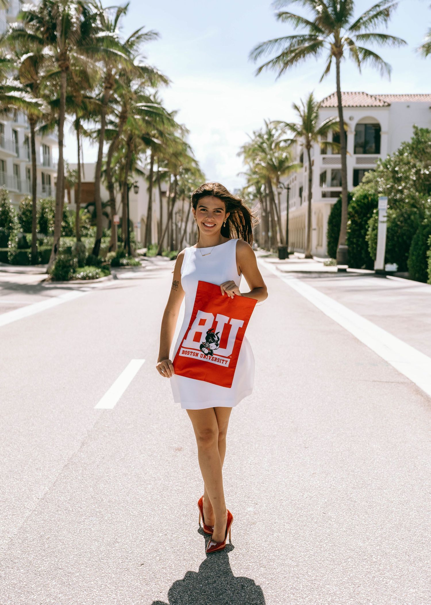 Young woman in white dress holding BU Boston University tote bag walking on palm tree-lined street on Worth Avenue Palm beach.