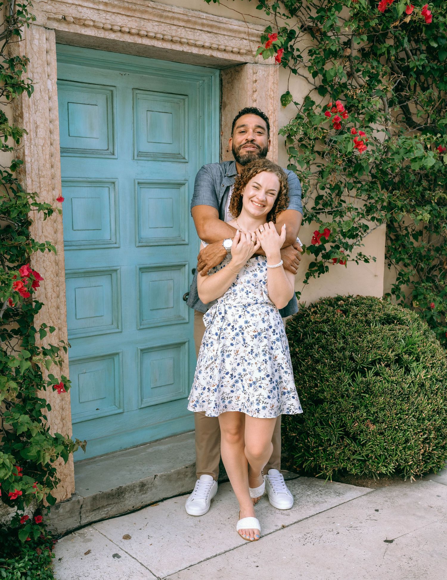 Couple smiling and holding a small dog in front of a turquoise door surrounded by red bougainvillea.Engagement photos on Worth Avenue