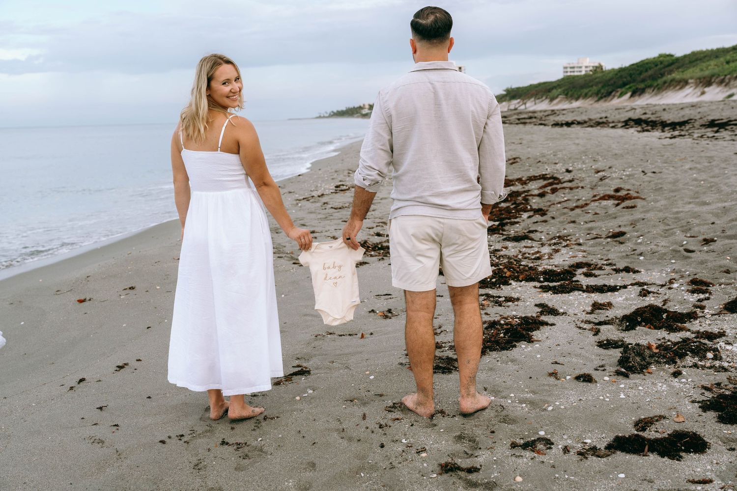 A couple in white outfits walks barefoot on a rocky beach, holding hands with their baby on a cloudy day.