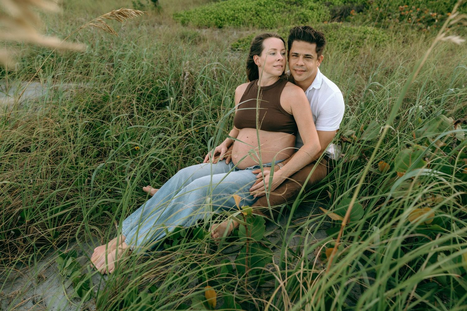 Pregnant couple sitting in tall beach grass, man embracing woman showing baby bump.