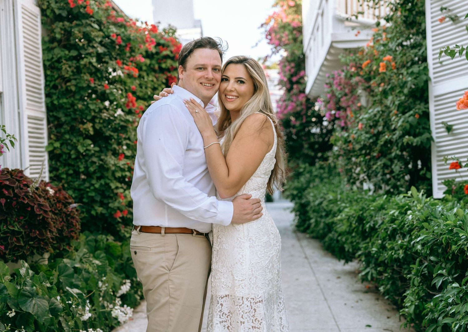 Couple embracing in a lush garden alley with colorful flowers, woman in white lace dress, man in white shirt.
