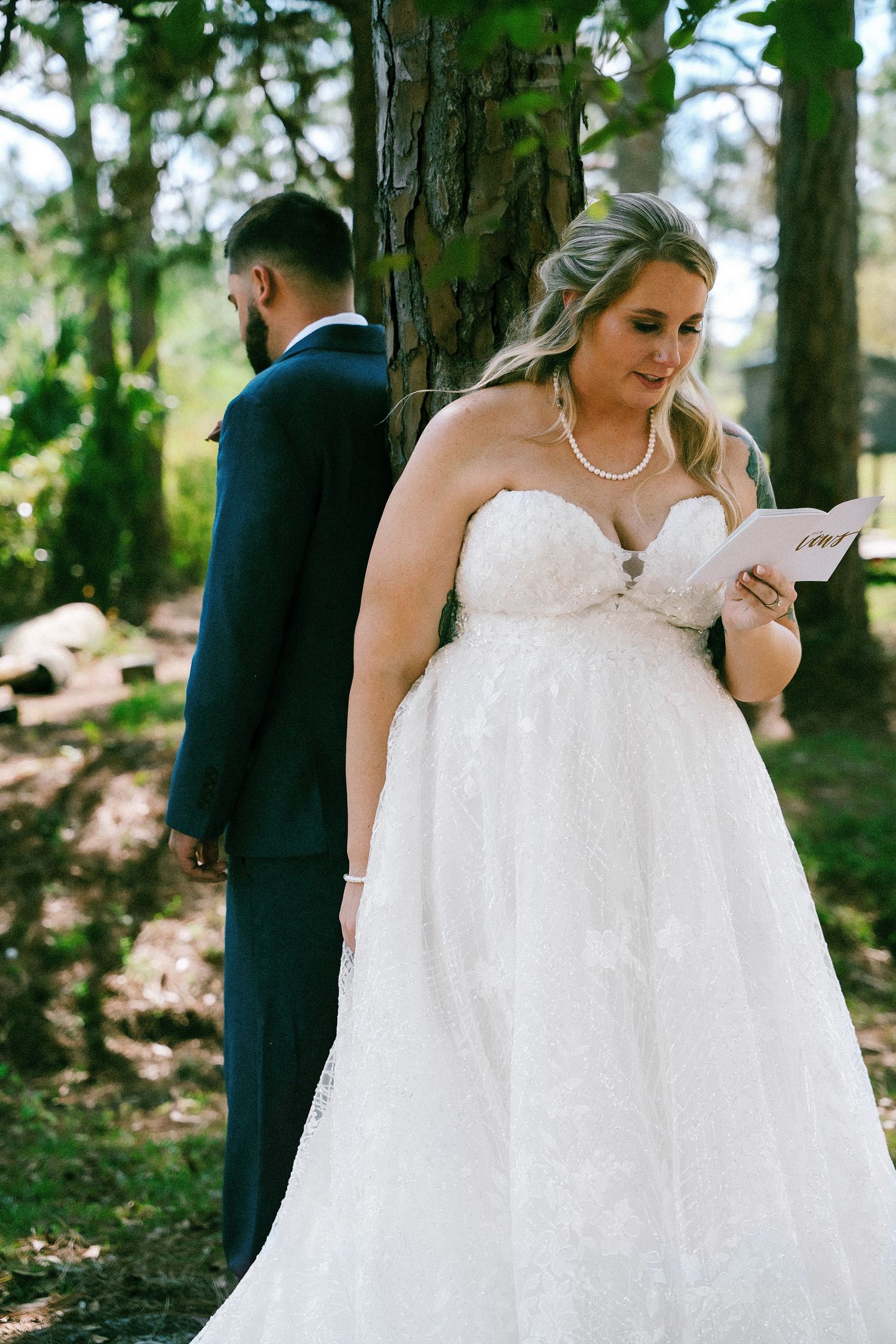 Bride in strapless lace ball gown reads vows from card while groom stands facing tree in forest wedding setting.