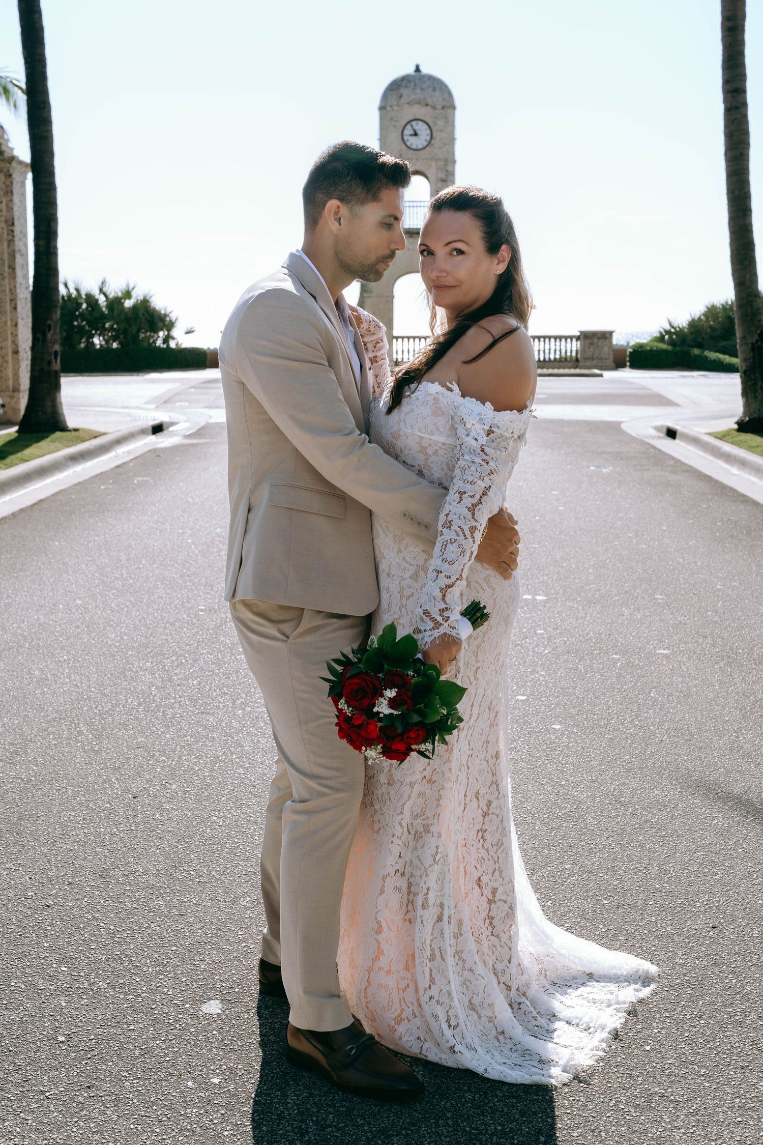 Bride in lace gown holding red roses embraces groom in tan suit near a clock tower on a sunny day.
