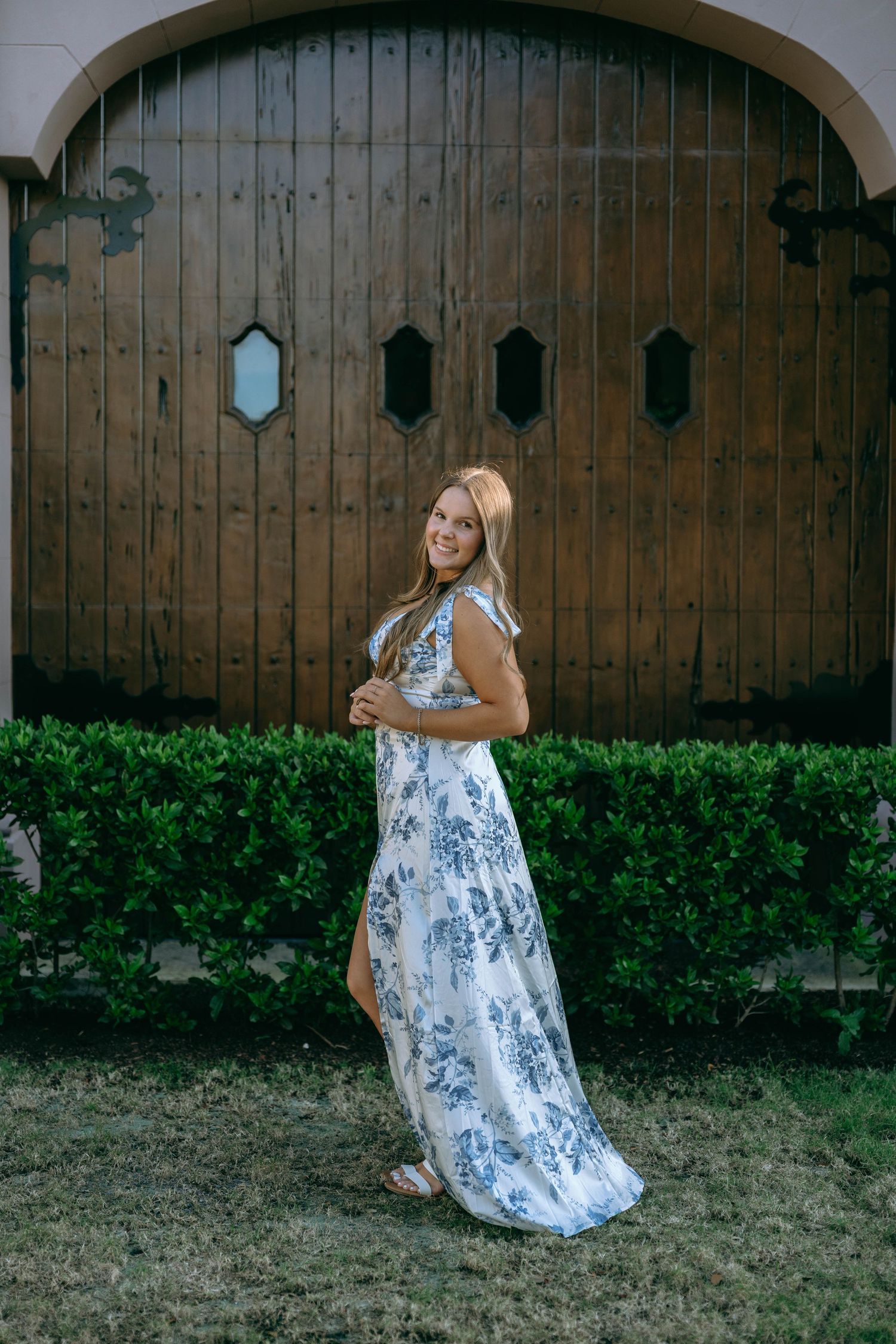 Smiling woman in blue floral maxi dress posing before rustic wooden doors with green hedges.