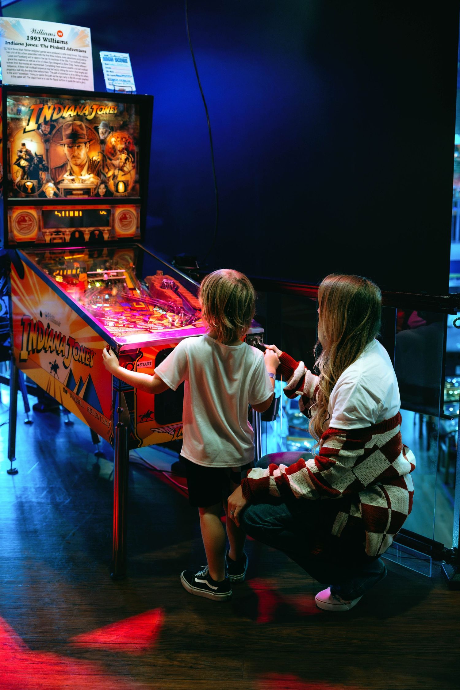 Child and woman playing Indiana Jones pinball machine together in a dimly lit arcade.