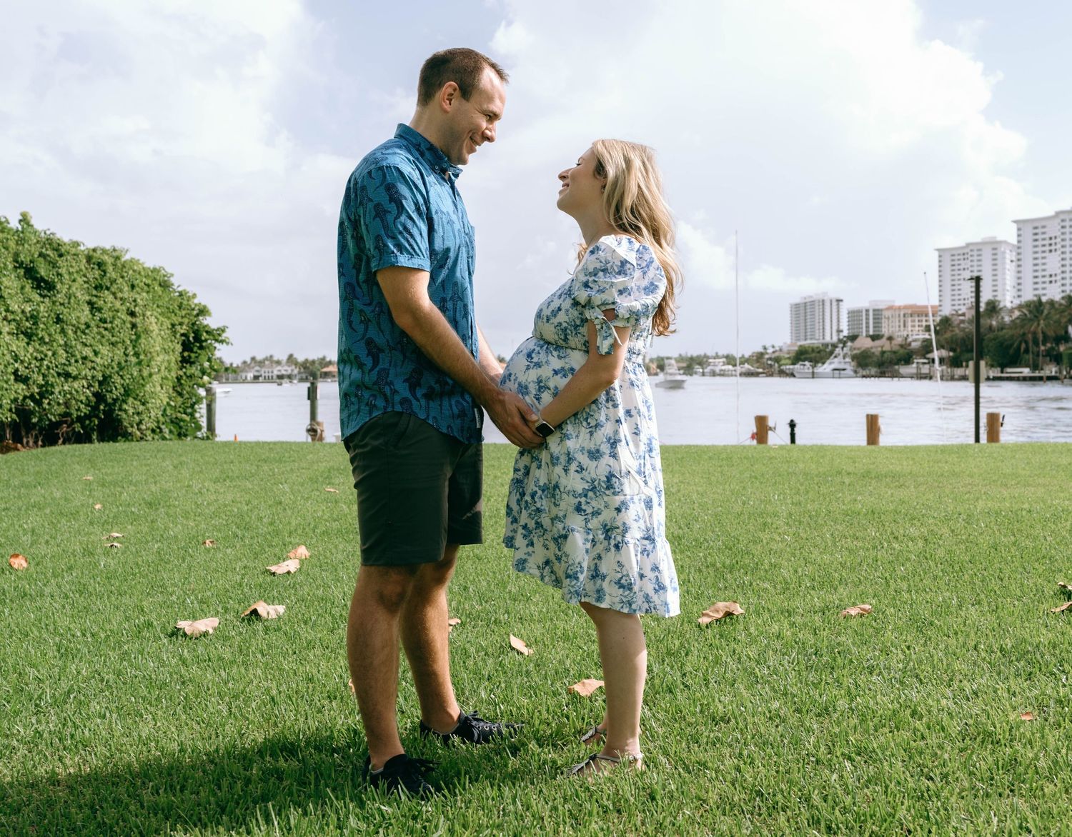Pregnant couple holding hands on green lawn by waterfront, woman in floral dress, man in blue Hawaiian shirt.