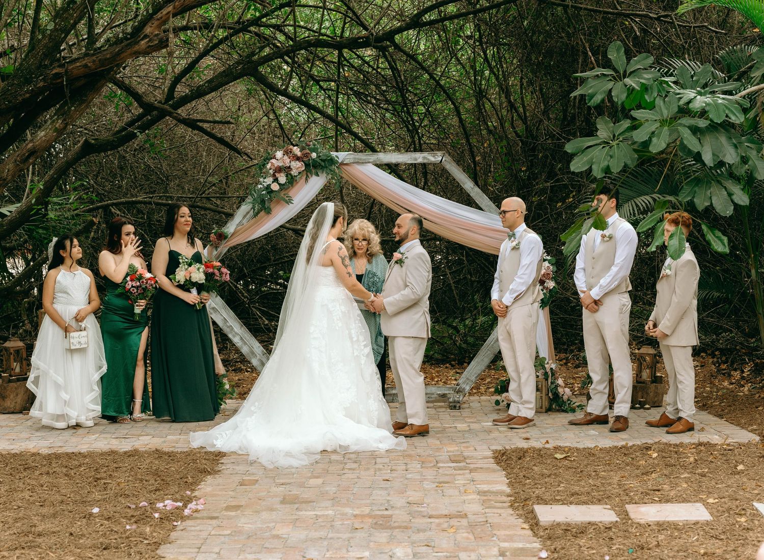 Wedding party celebrates on stone path under arching trees with bridesmaids in green and groomsmen in beige suits.