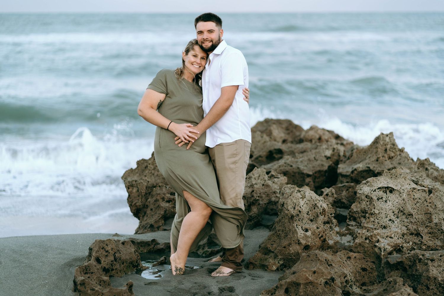 A couple embraces on rocky coastal formations with ocean waves in the background.