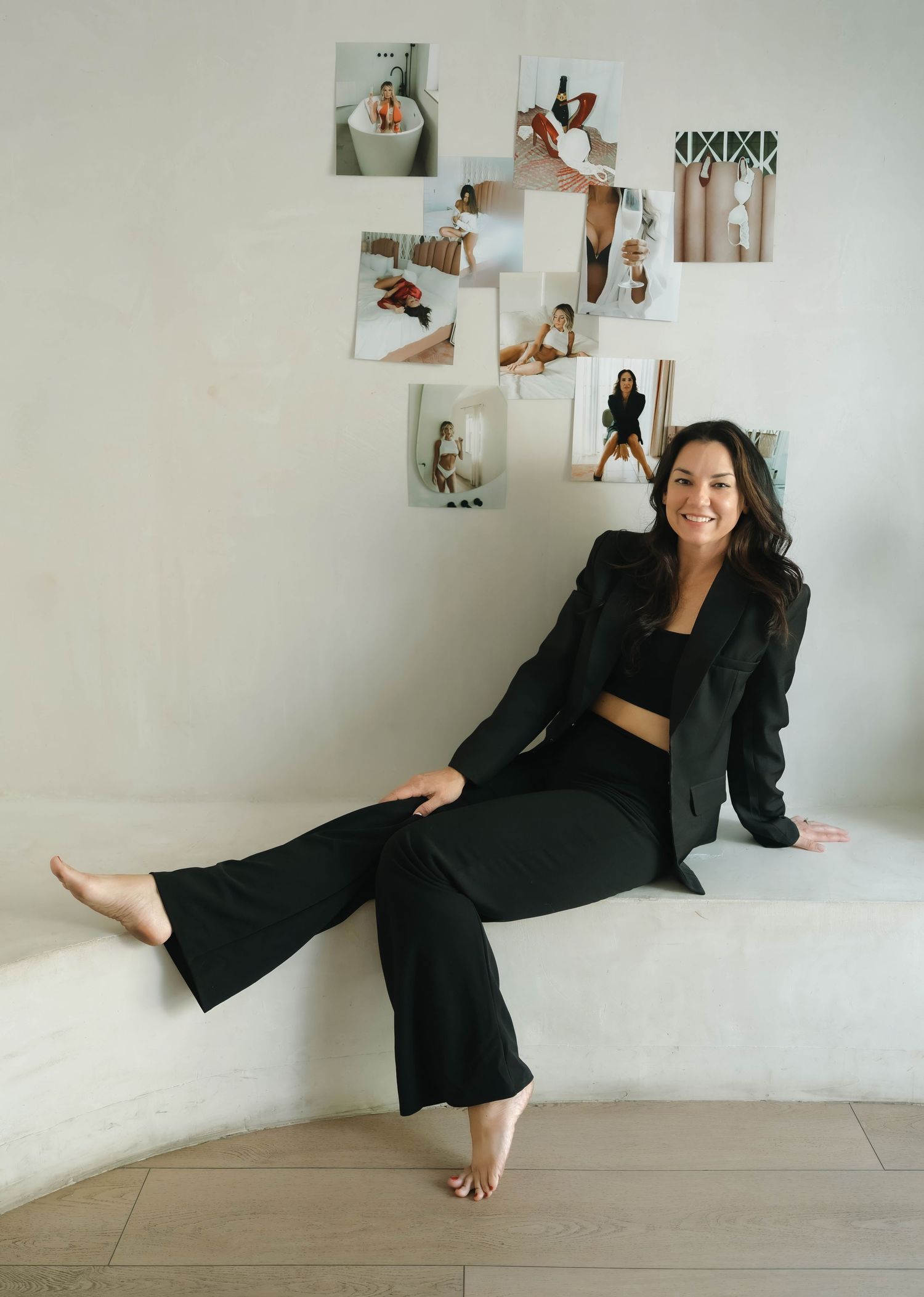 A woman in black business attire sits casually against a white wall with photos displayed.