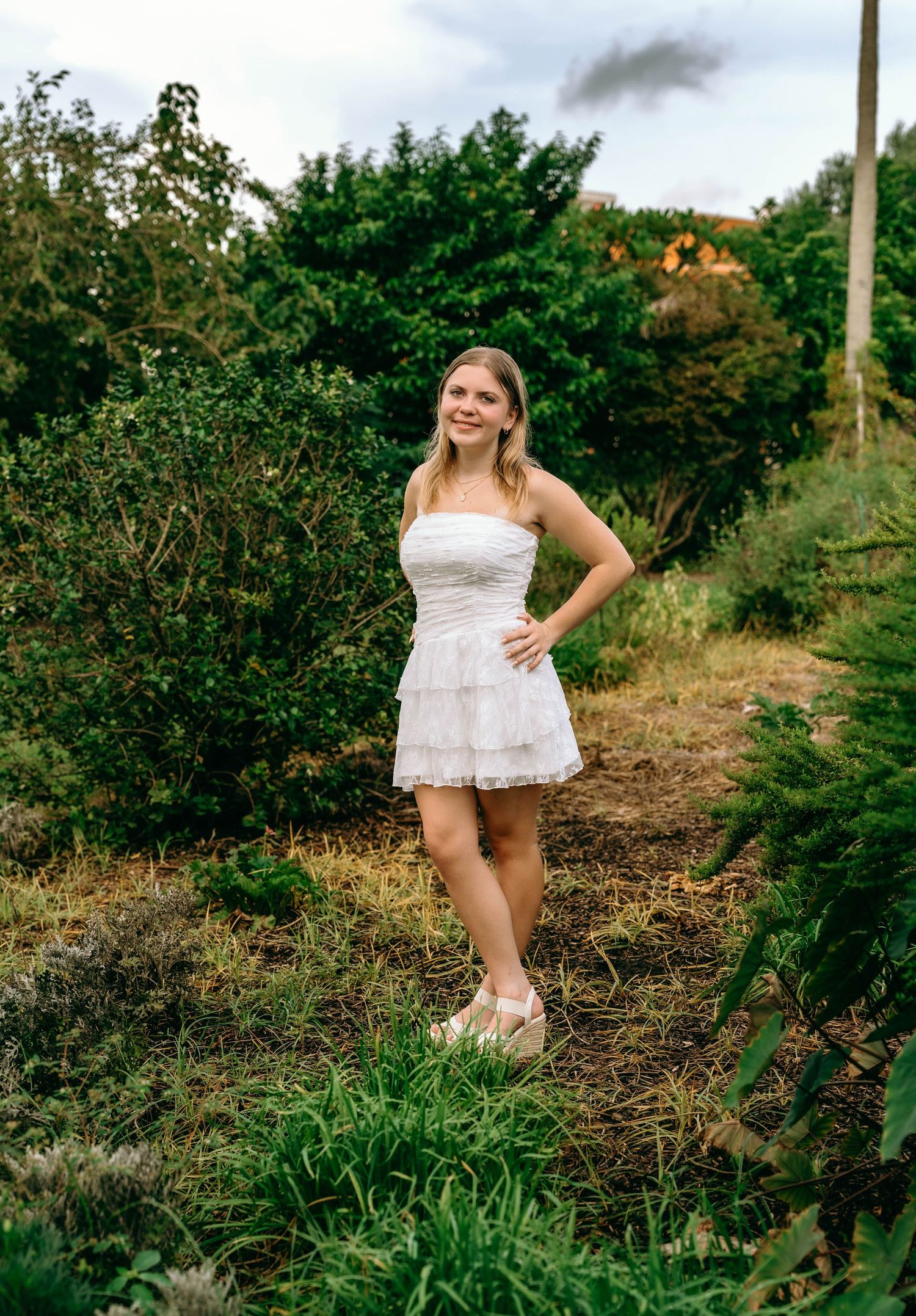 Smiling young woman in white ruffled strapless dress posing among garden shrubs outdoors.
