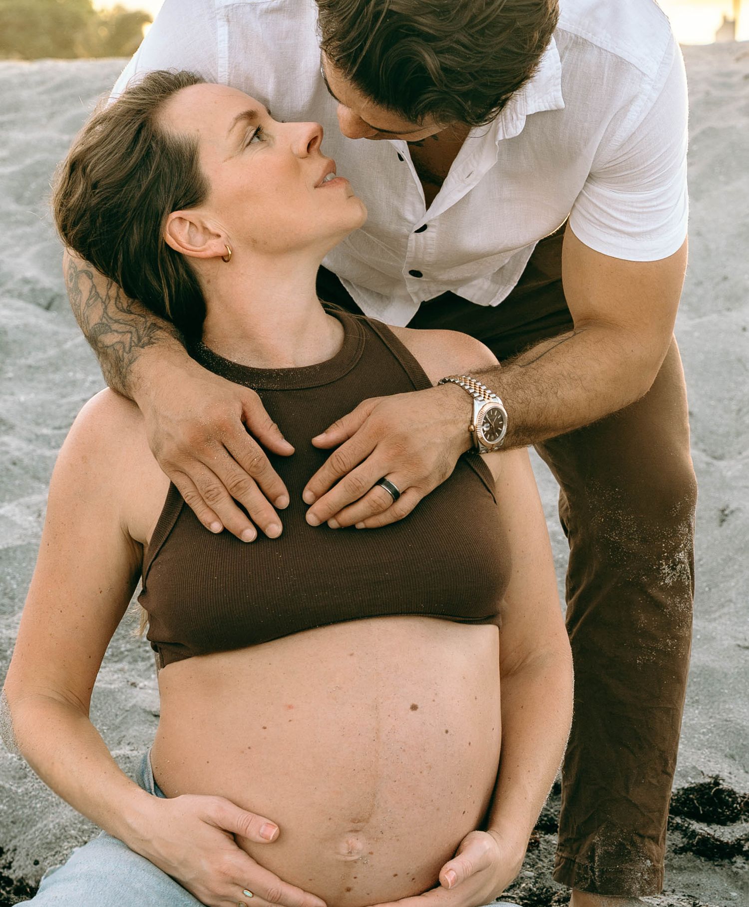 A pregnant woman sits on a beach while her partner lovingly holds her baby bump during a maternity photo shoot.
