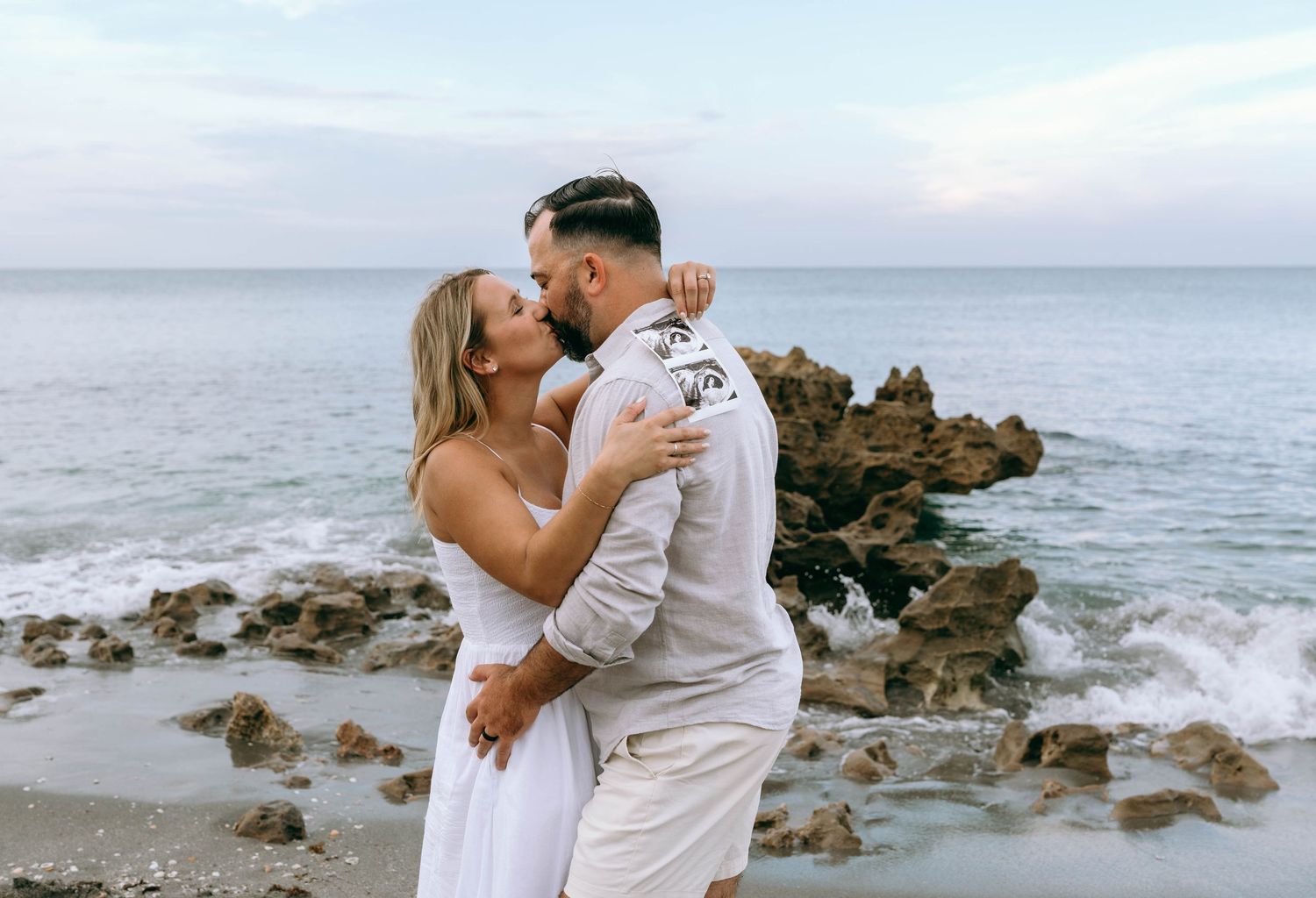 Couple sharing a romantic kiss on a rocky beach with ocean waves, woman in white dress, man in beige outfit.