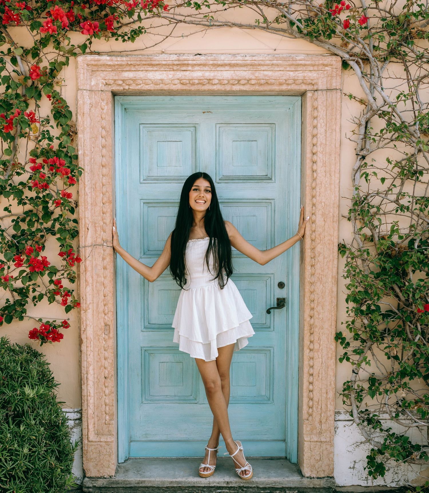 Young woman in white dress posing by a turquoise door surrounded by red bougainvillea flowers on Worth Avenue Palm beach.