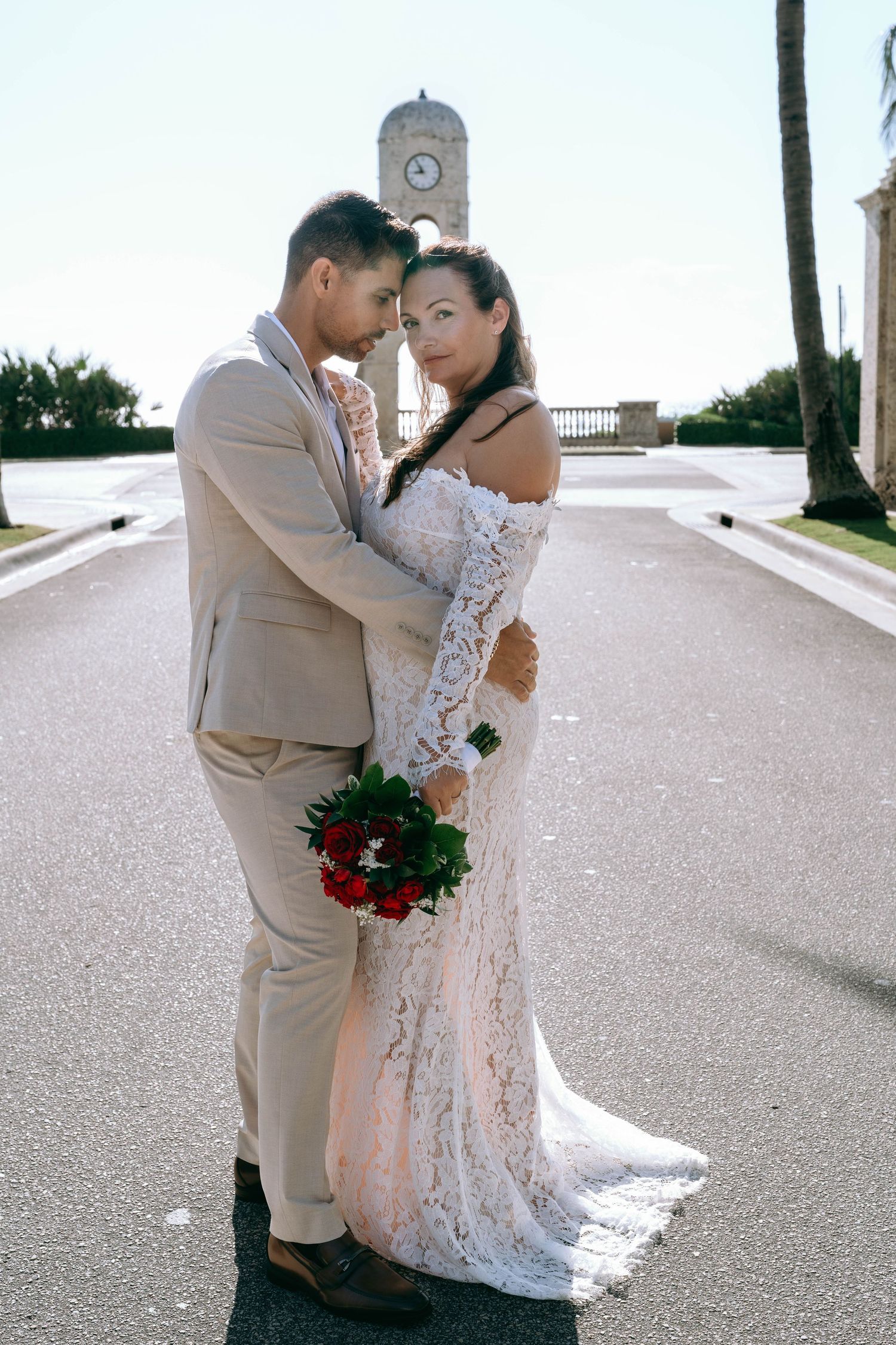 Bride and groom embracing on a sunlit path with a clock tower backdrop, bride holding red bouquet in white lace gown.