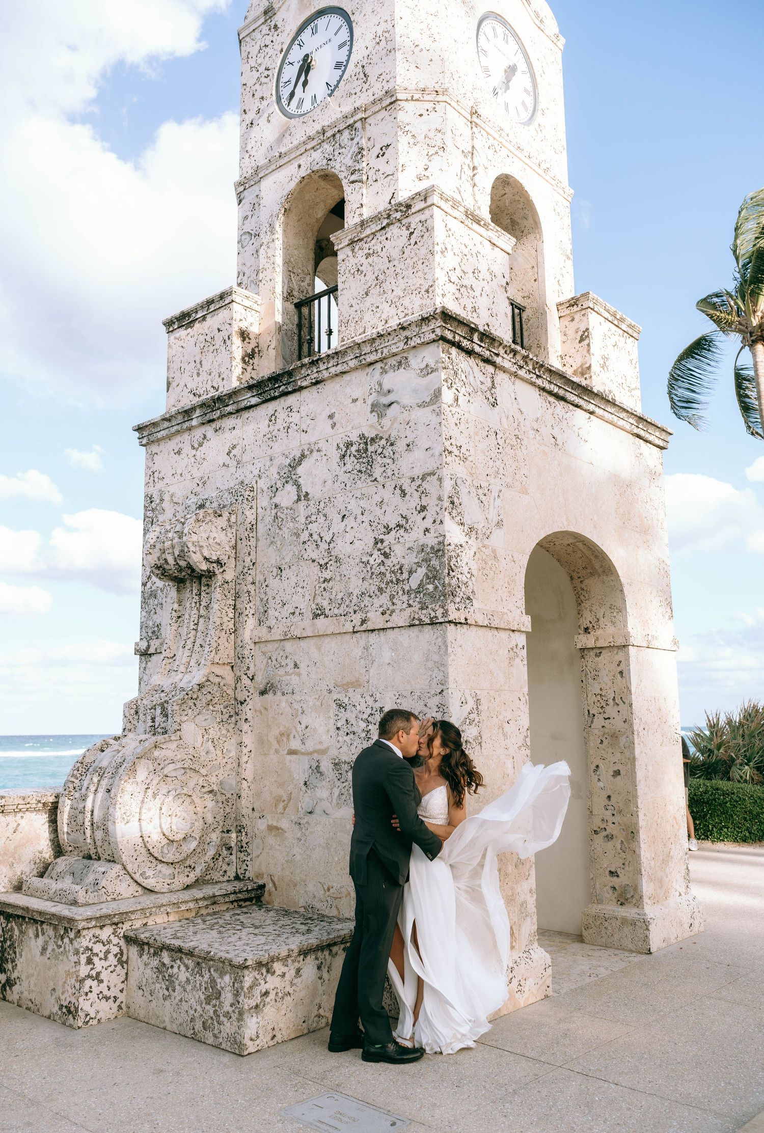 Couple shares a kiss beneath a historic clock tower with ocean views on their wedding day.