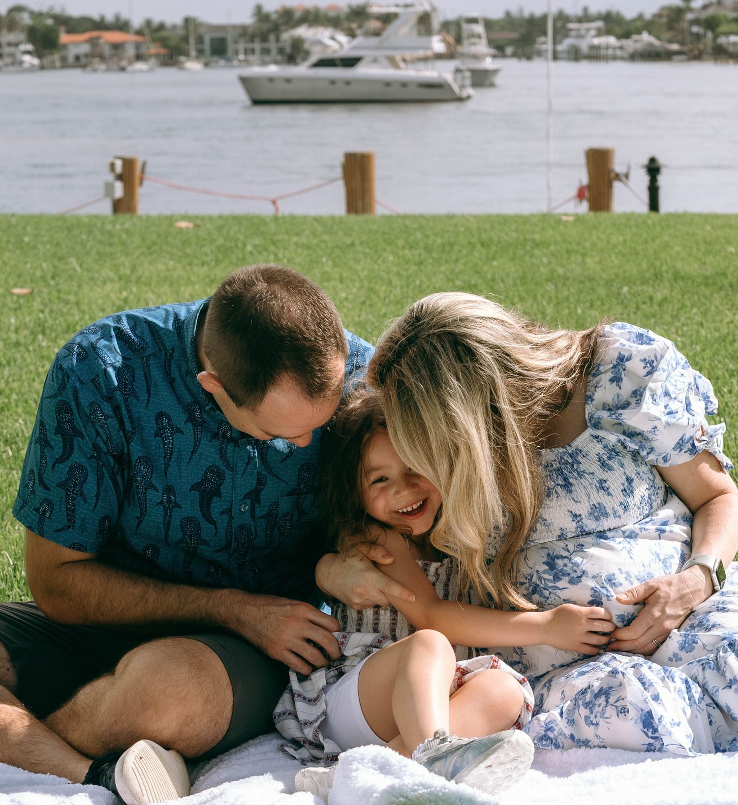 A happy family of three sits on a blanket by a waterfront park, sharing a joyful embrace on a sunny day.