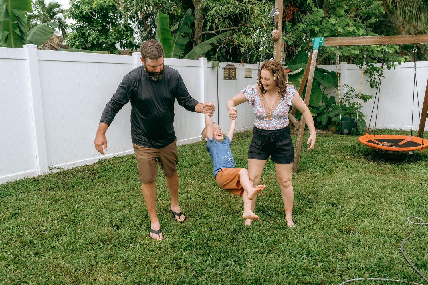 Family playing in backyard, parents swinging toddler on green lawn near white fence and wooden swing set.