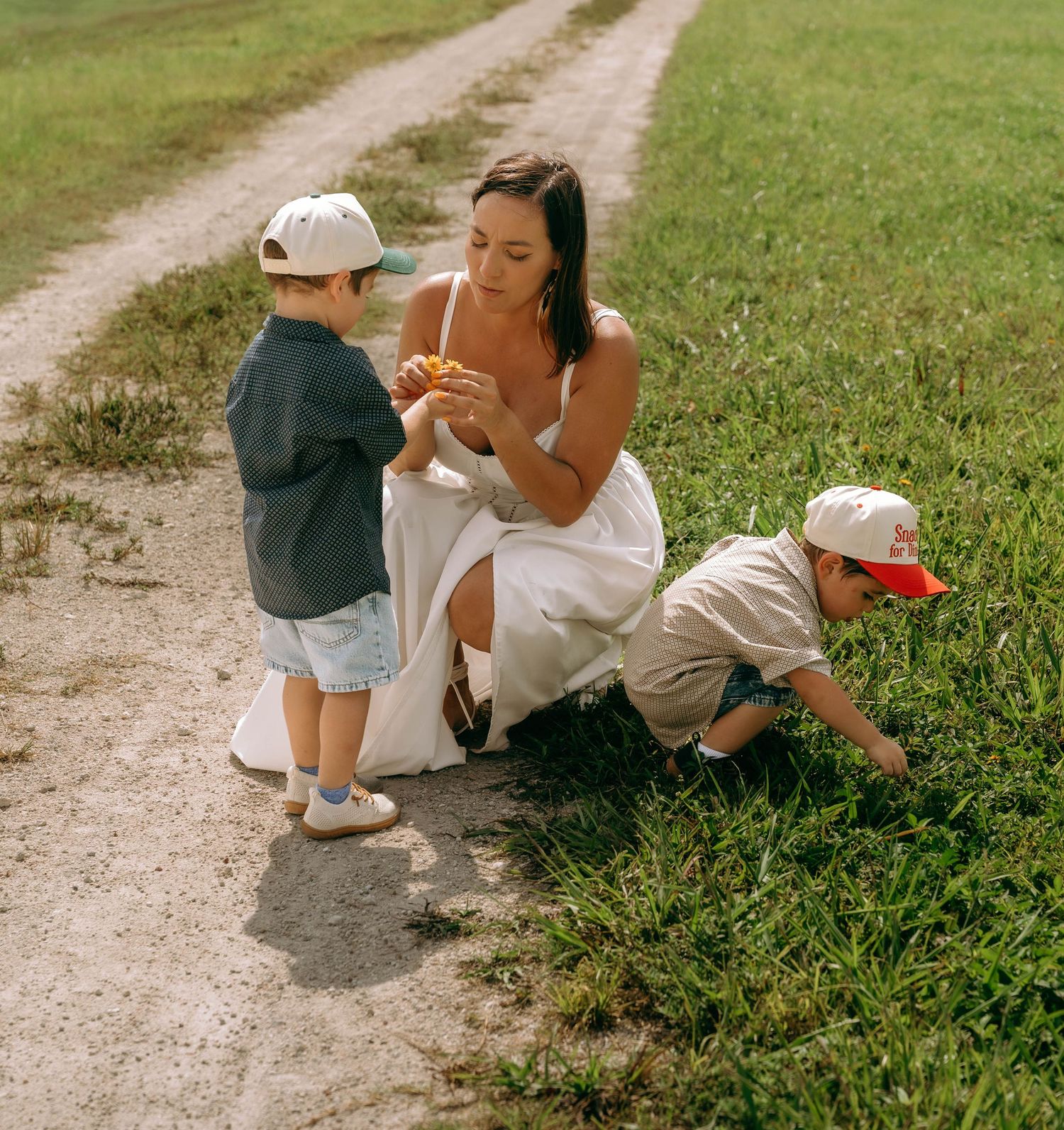 A woman in a white dress crouches on a dirt path with two young boys in baseball caps exploring the green grass.