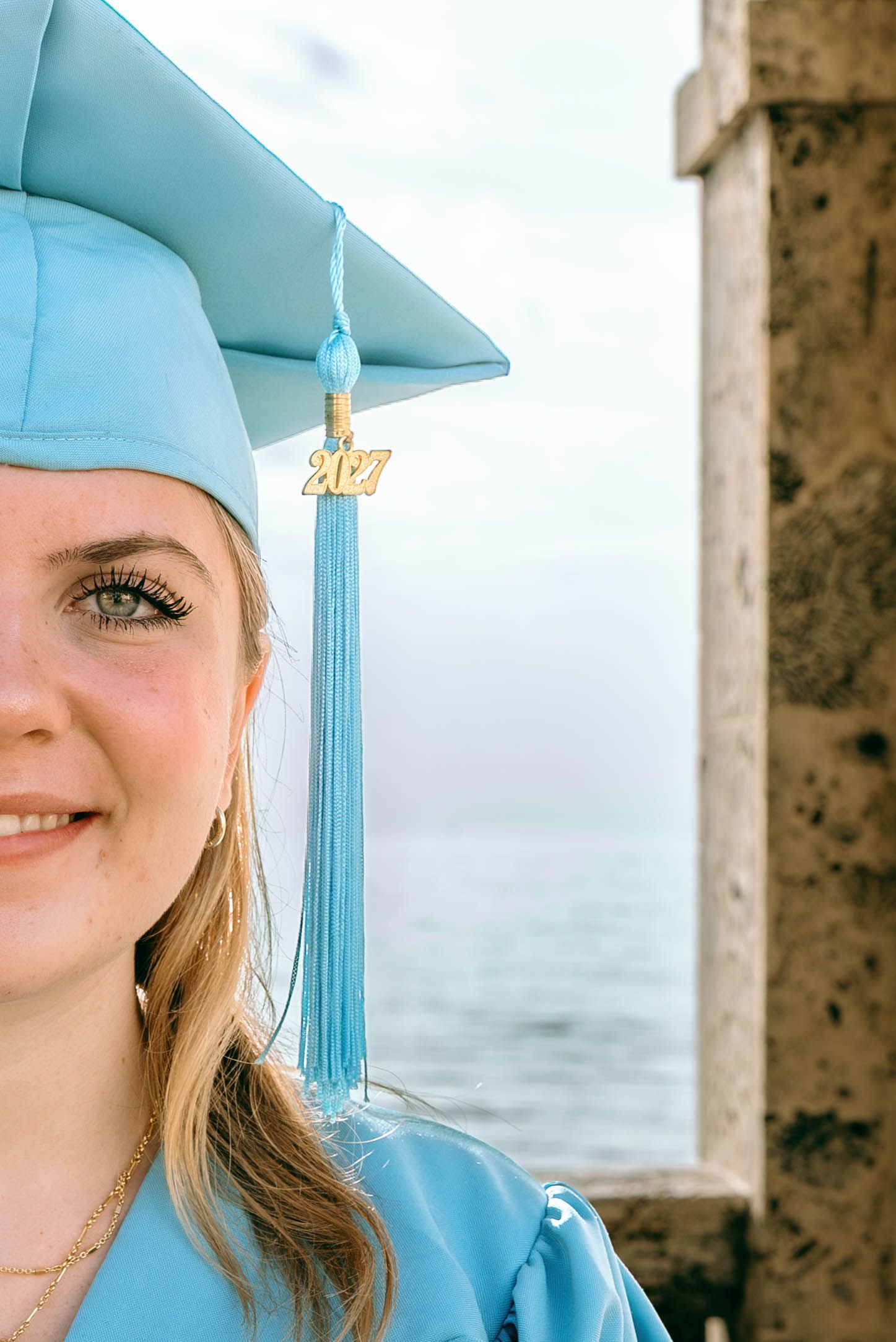 Smiling young woman in light blue 2027 graduation cap and gown posing outdoors near the ocean.