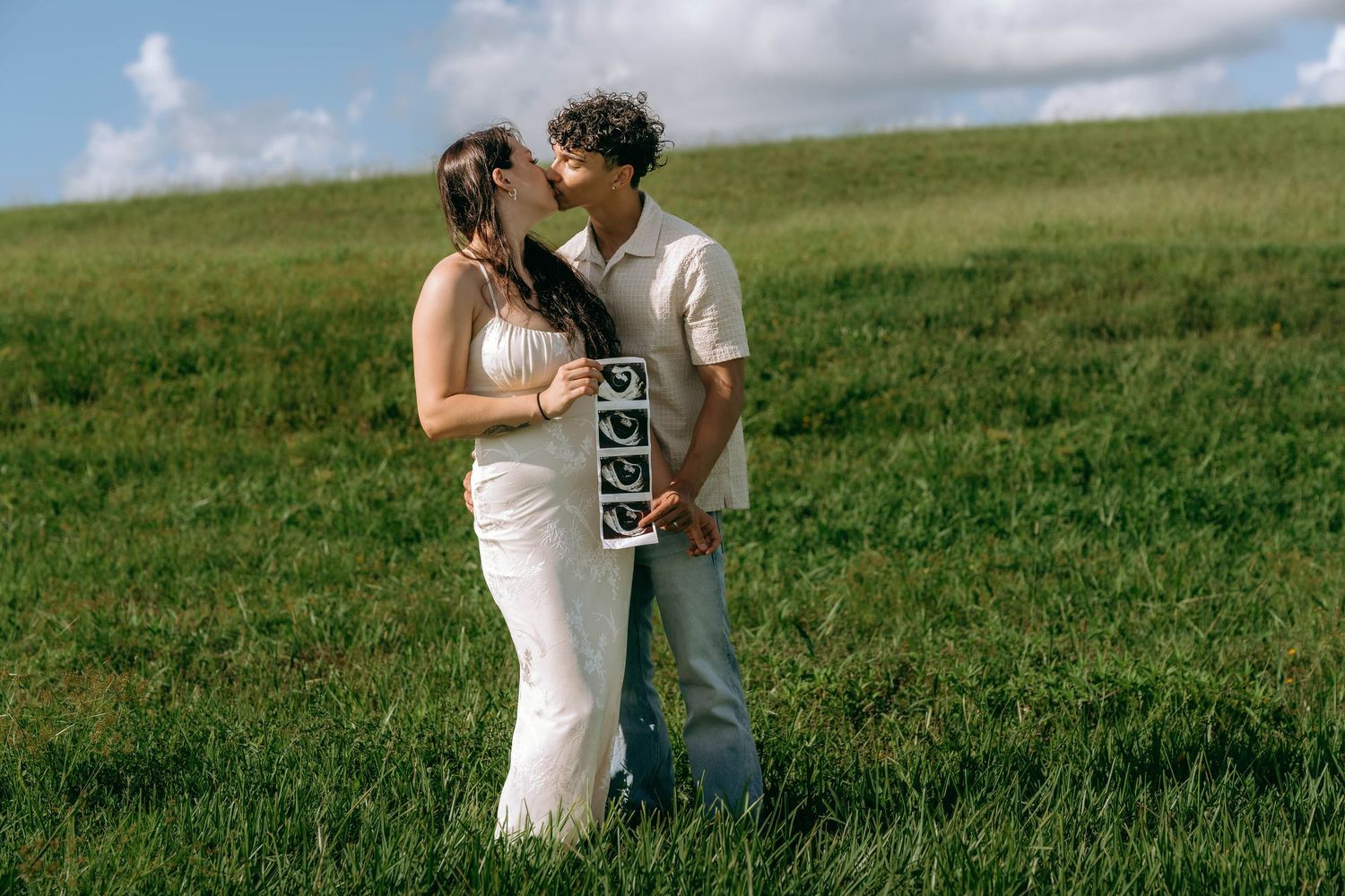 Couple kissing in green field, woman in white dress holding ultrasound photos, pregnancy announcement outdoors.