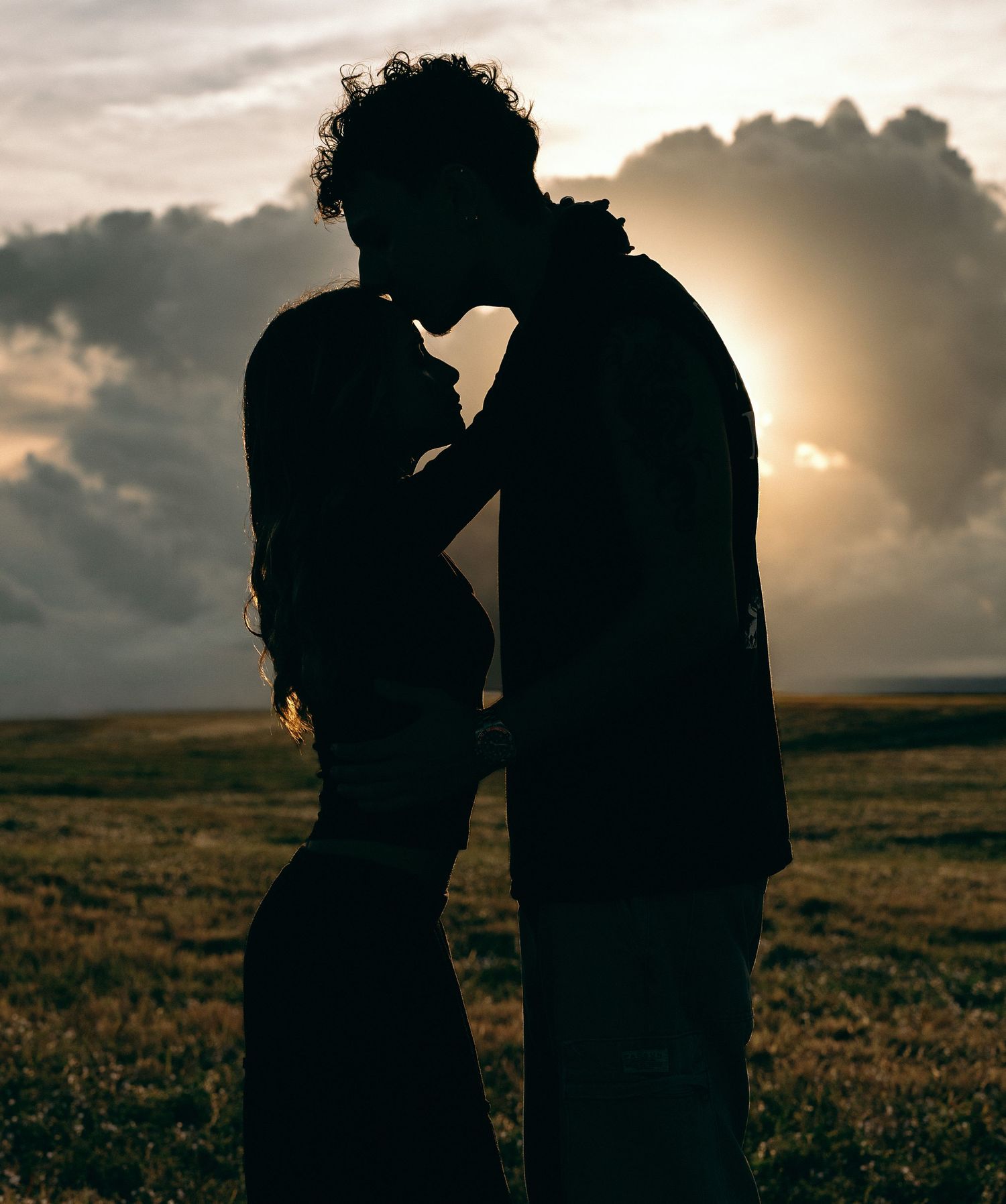 Silhouettes embrace against a dramatic sunset sky over rolling hills.