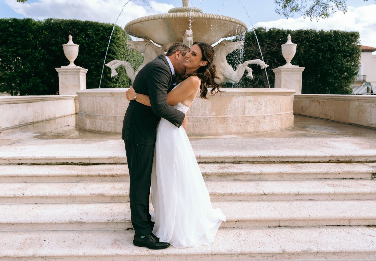 Newlywed couple sharing a romantic kiss in front of an elegant stone fountain on their wedding day.