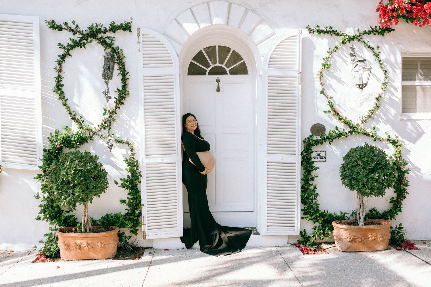 Pregnant woman in elegant black gown posing by white arched door with lush green ivy topiaries. Worth Avenue, Palm Beach, FL