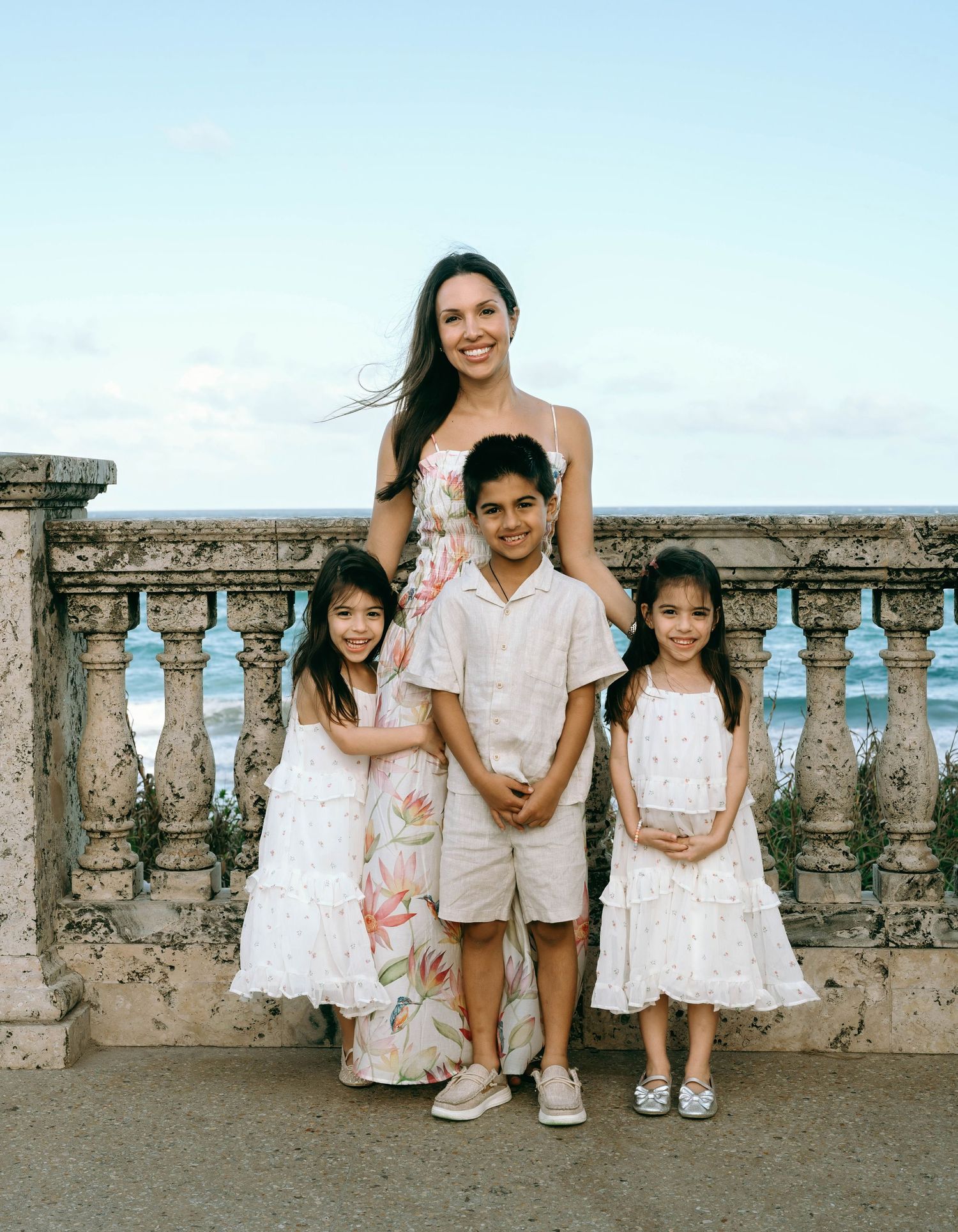 A mother poses with her three children on a seaside balustrade terrace, all dressed in white and cream outfits. Worth Avenue, Palm Beach, FL
