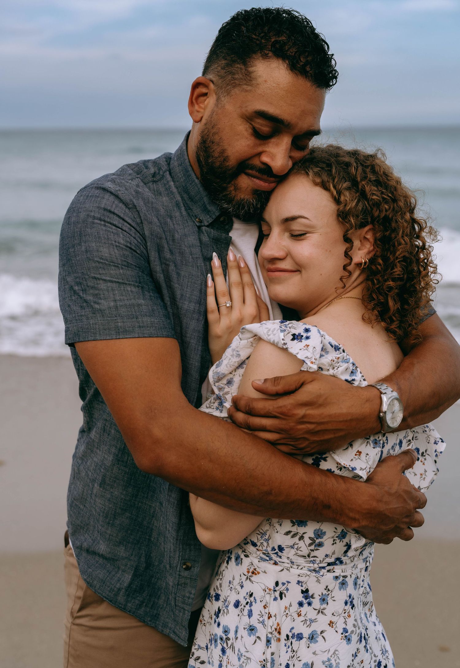 Tender couple embracing on a beach at dusk, woman in floral dress smiles peacefully in mans arm on Worth Avenue Palm beach.