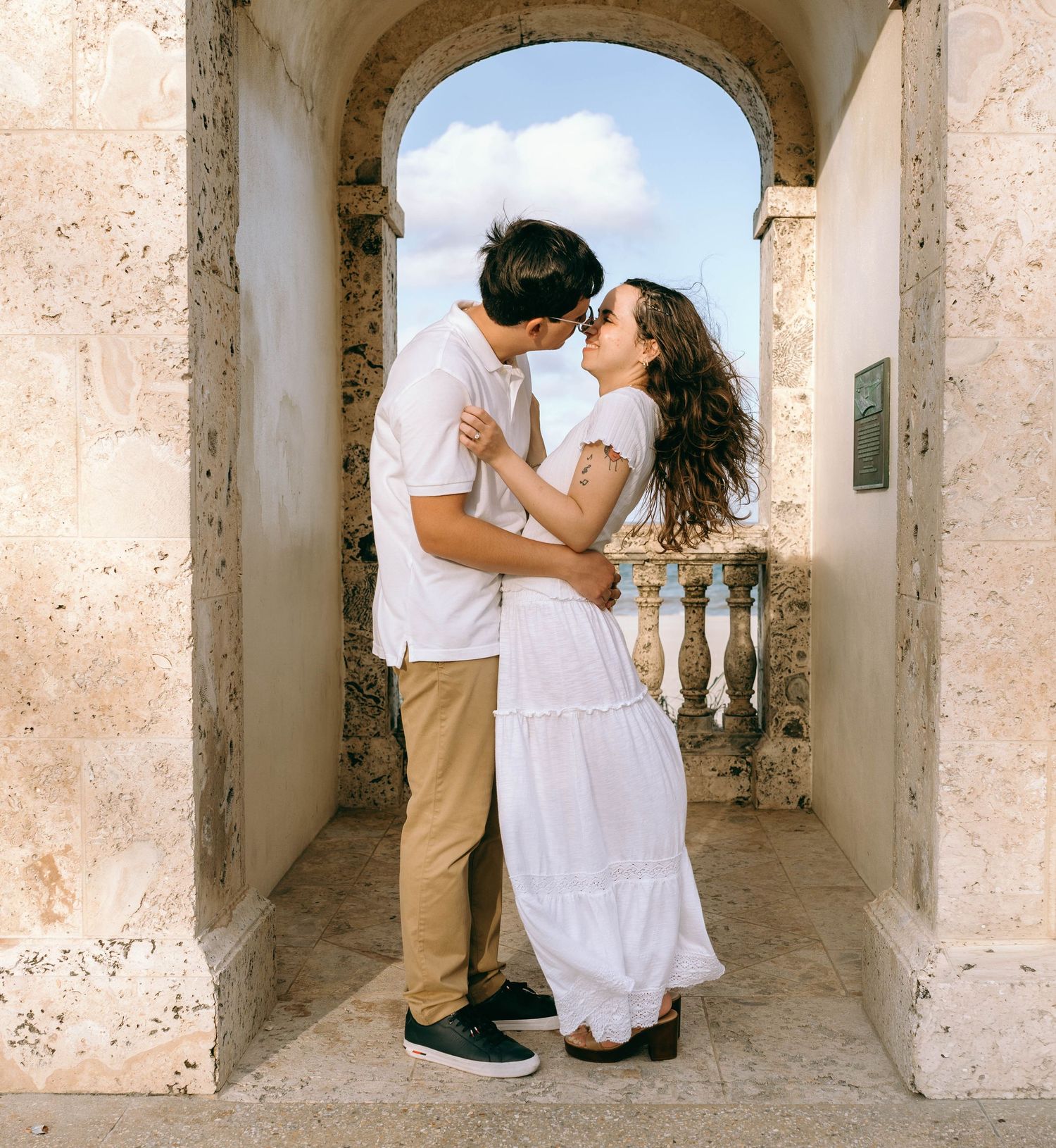 Young couple sharing a kiss while embracing under a stone archway with a blue sky backdrop. Engagement photos on Worth Avenue in Palm Beach