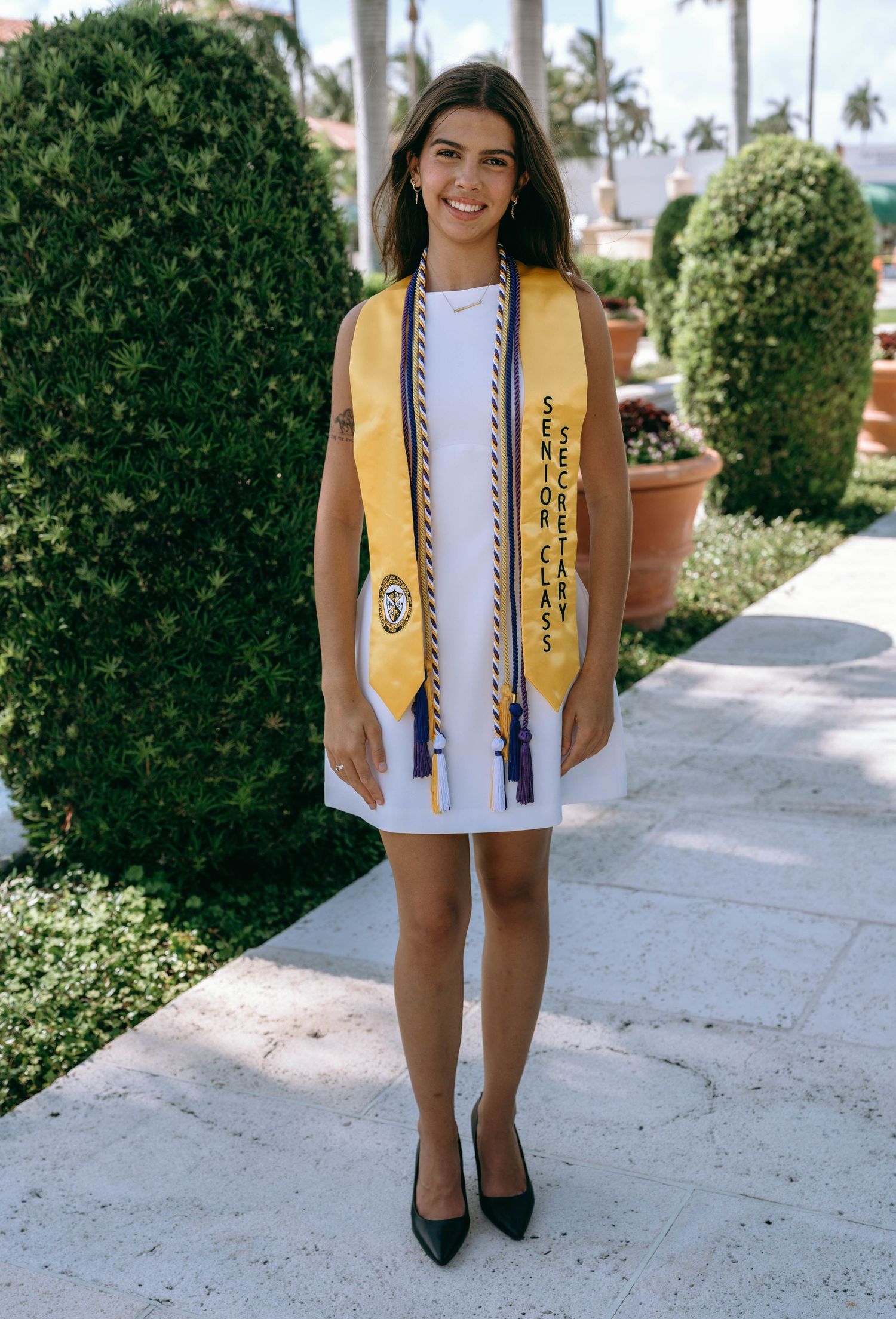 Smiling graduate in white dress with yellow senior class stole and honor cords on garden path on Worth Avenue Palm beach.