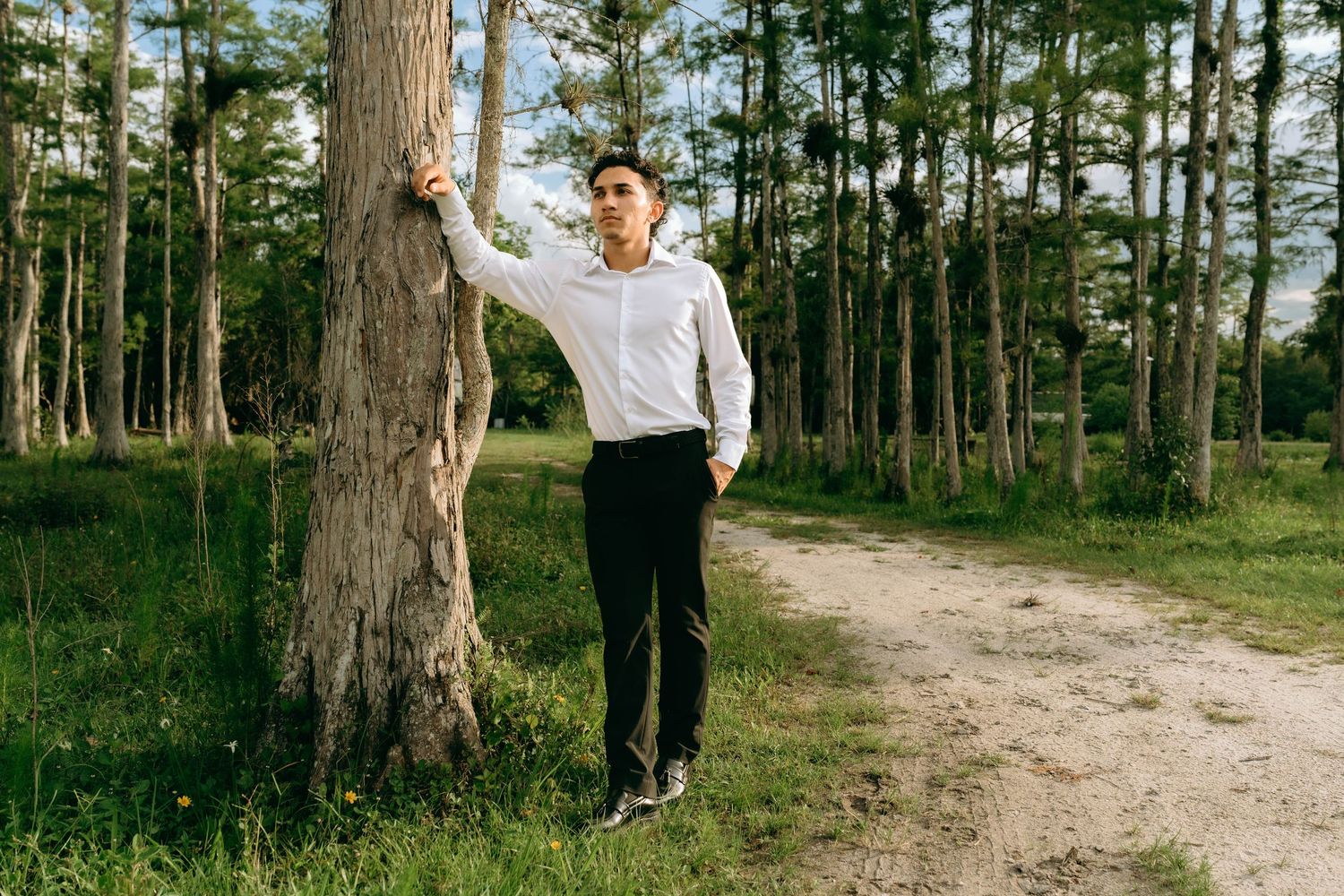Young man in white shirt and black pants leaning against a tree on a forest dirt path. Senior Photos at Colt Farms in Boynton Beach, FL