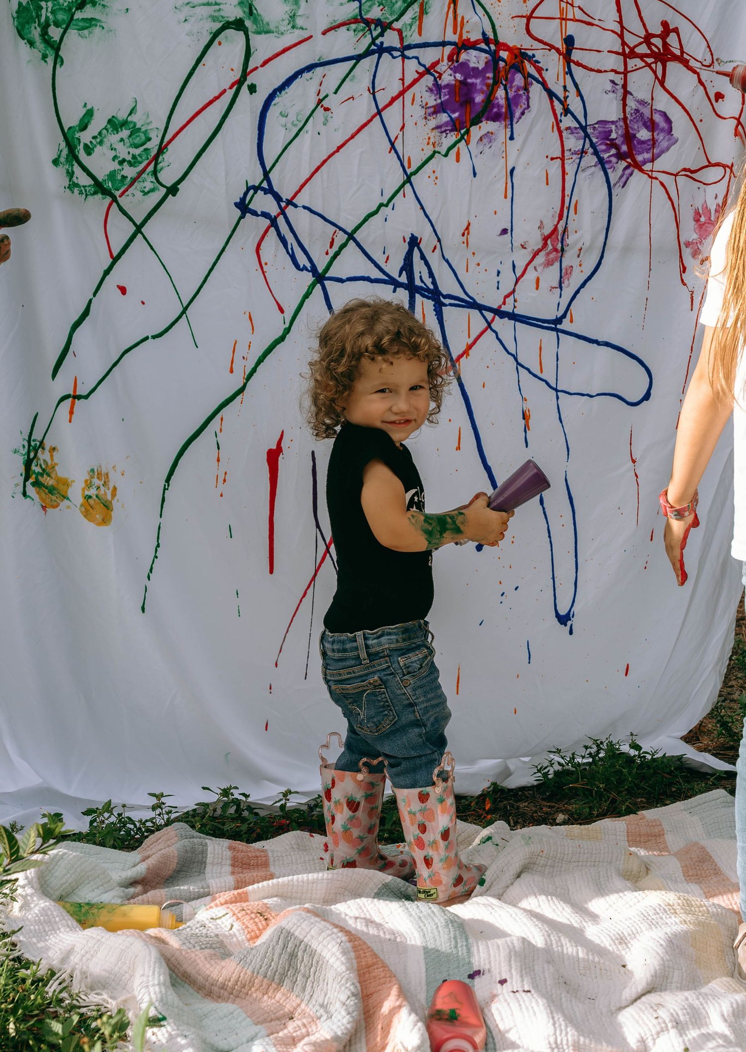 A curly-haired toddler in rain boots holds a paintbrush beside a colorful painted white canvas outdoors.