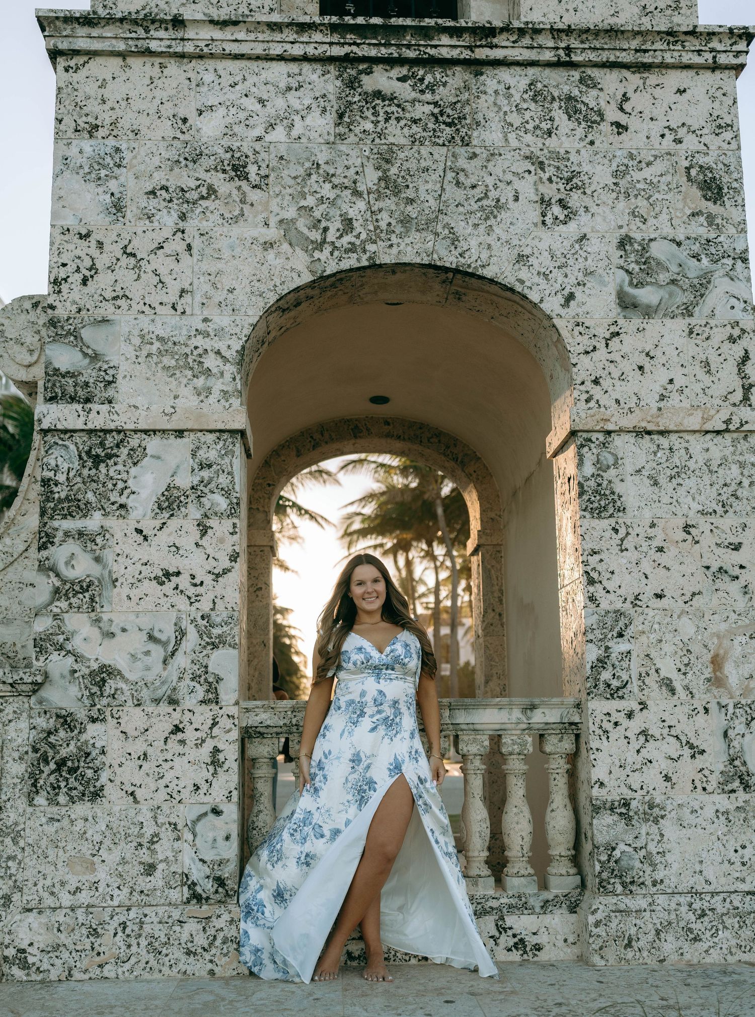 Woman in blue floral slit maxi dress standing in a weathered stone archway at sunset.