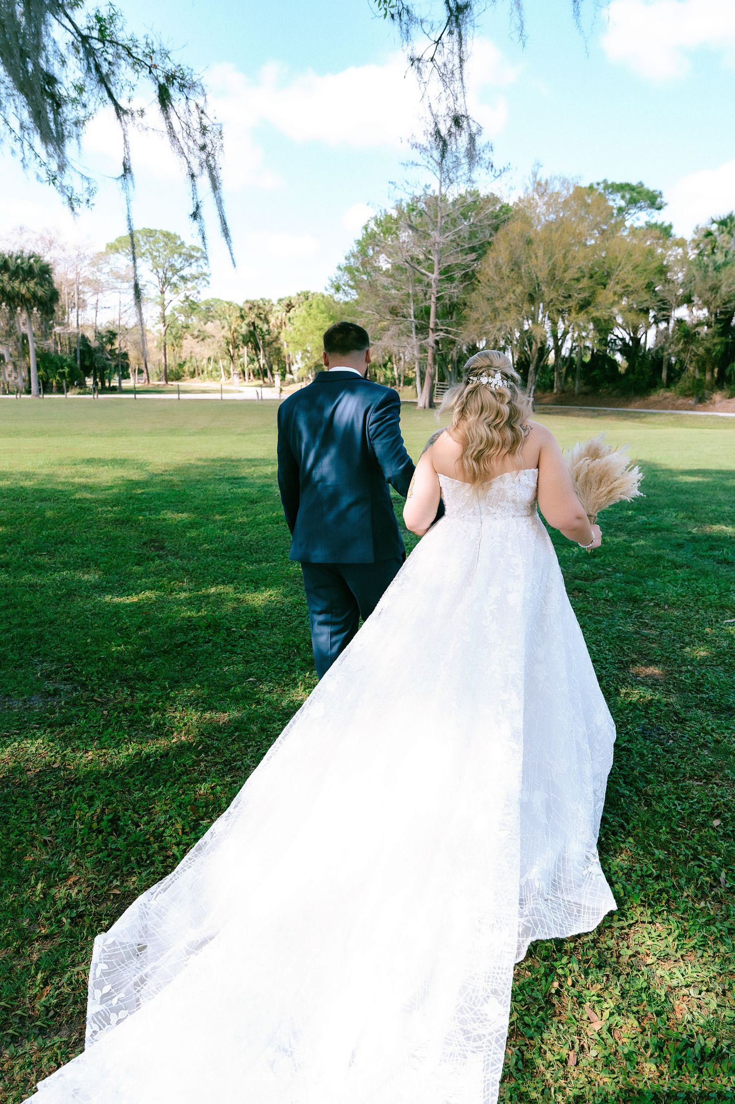 Bride and groom walk hand in hand across green lawn, bride's long lace train trailing behind in outdoor wedding.