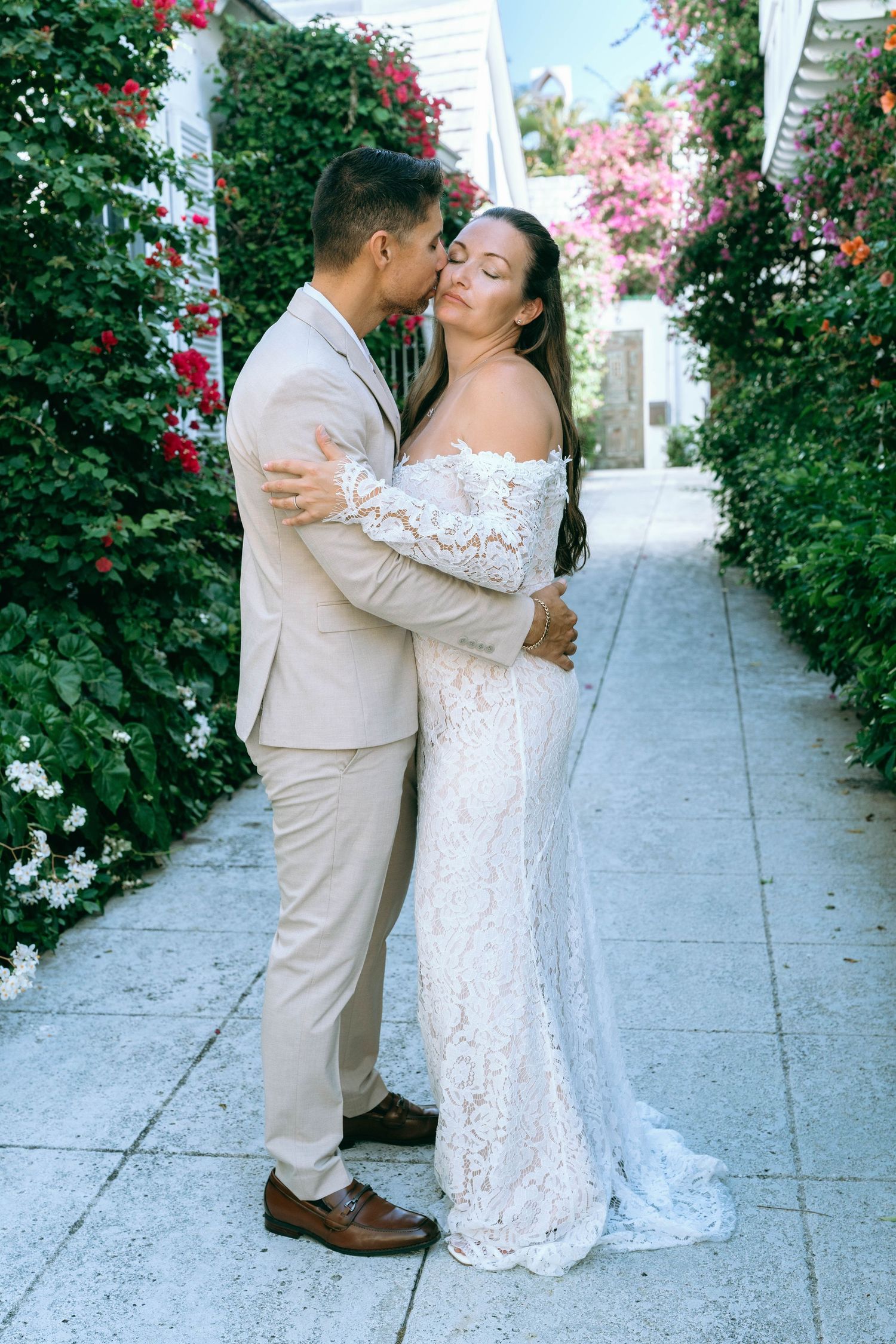 Bride and groom embracing in a floral garden pathway, bride in lace off-shoulder gown, groom in beige suit.
