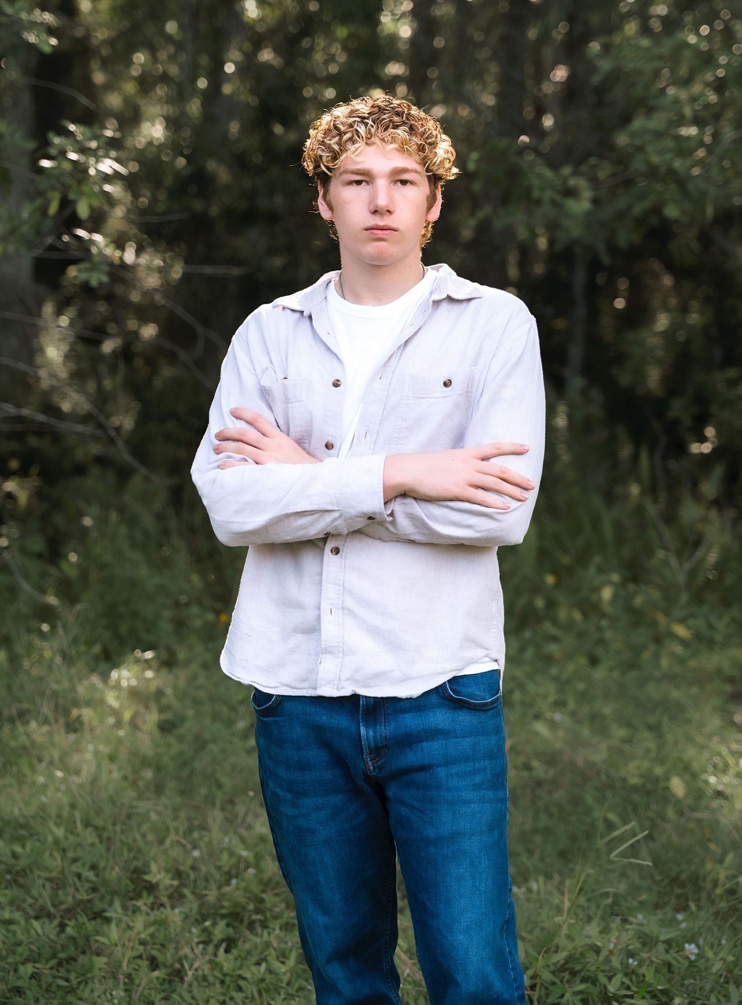 A young person in white shirt and jeans stands with crossed arms in a wooded setting.