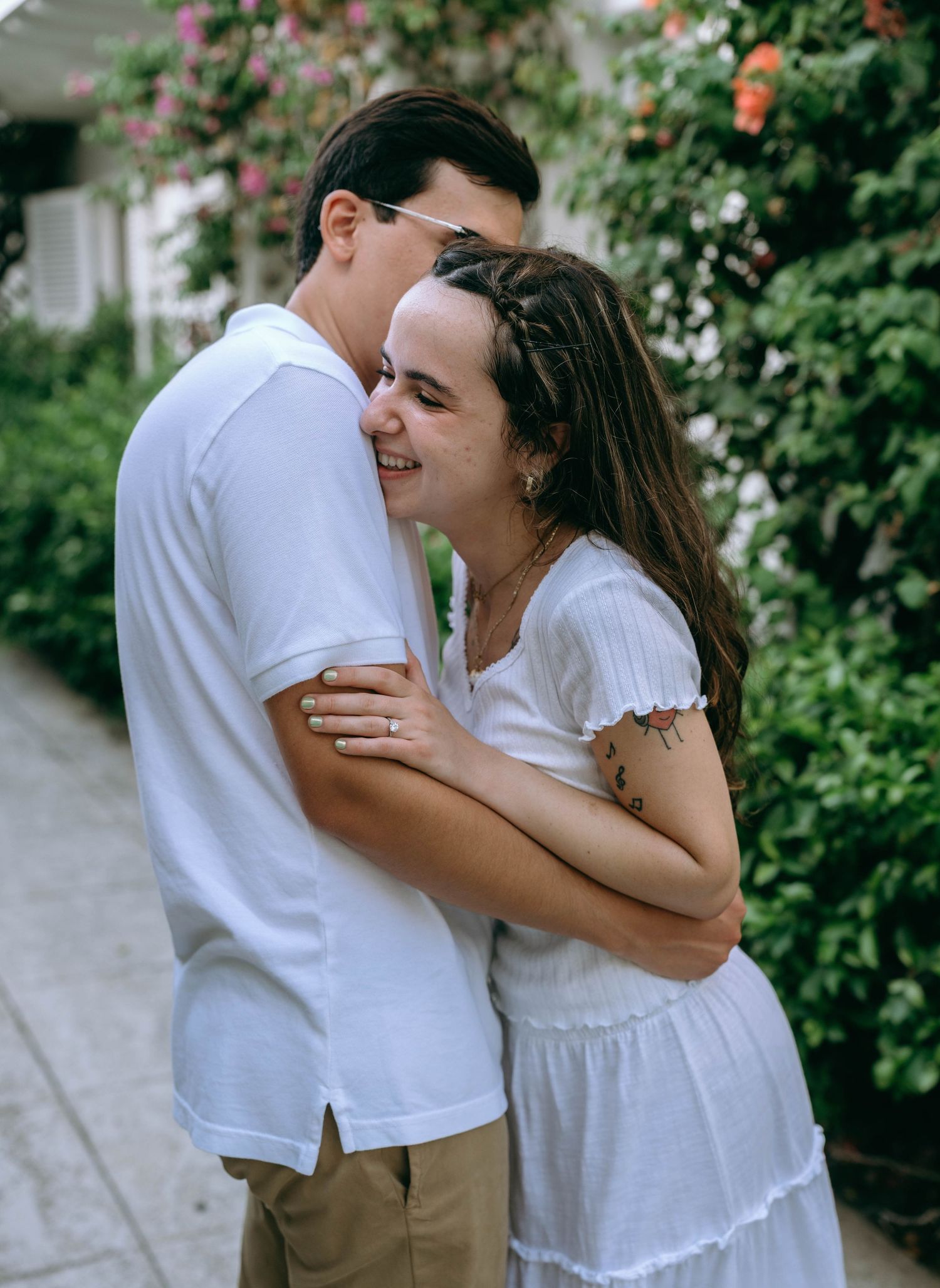 A young couple in white outfits embrace and smile in a lush garden pathway with flowering vines. Engagement photos on Worth Avenue