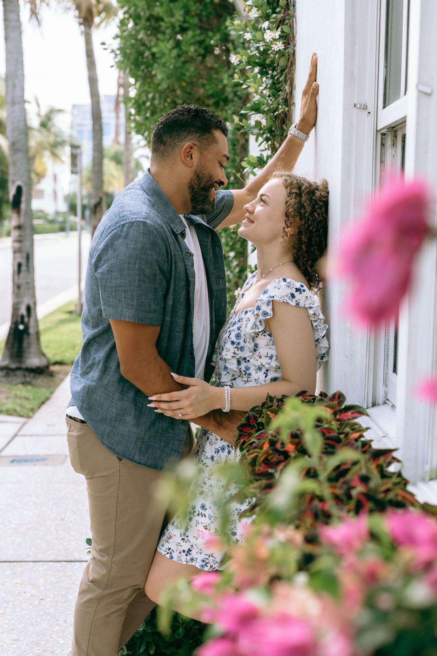 Romantic couple in floral dress and blue shirt leaning close together by a white wall with pink flowers. Engagement photos on Worth Avenue