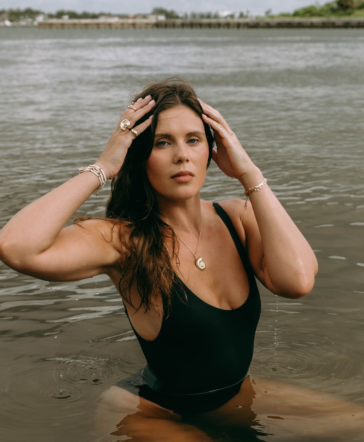Woman in black swimsuit wearing gold jewelry poses in shallow water near a shoreline.