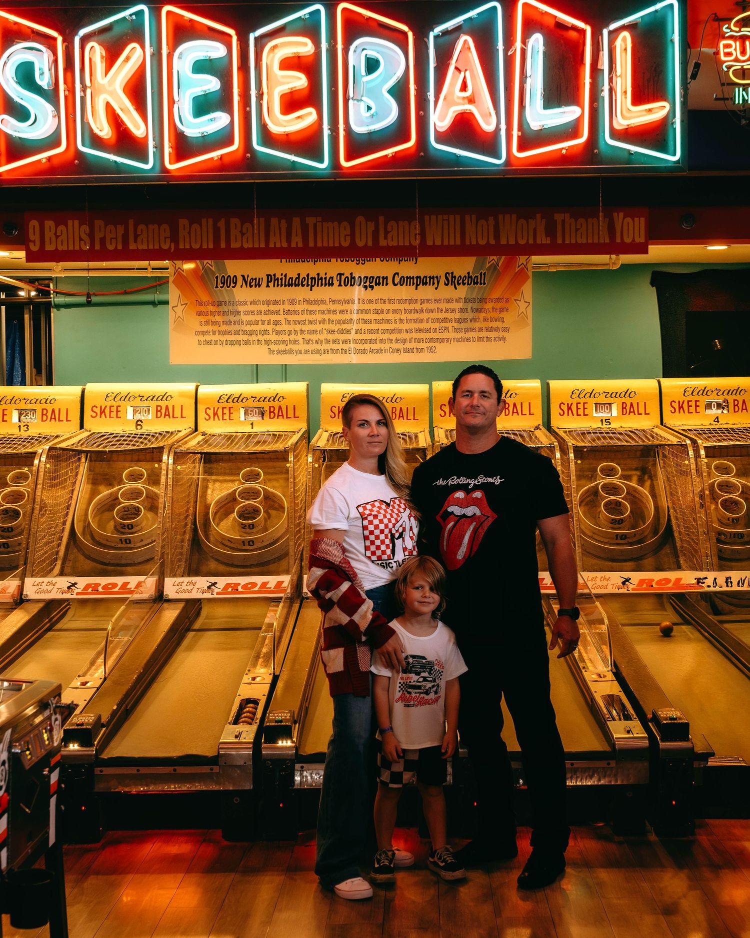 Family of three posing in front of Skee-Ball lanes at an arcade, with neon signs and rolling balls visible.