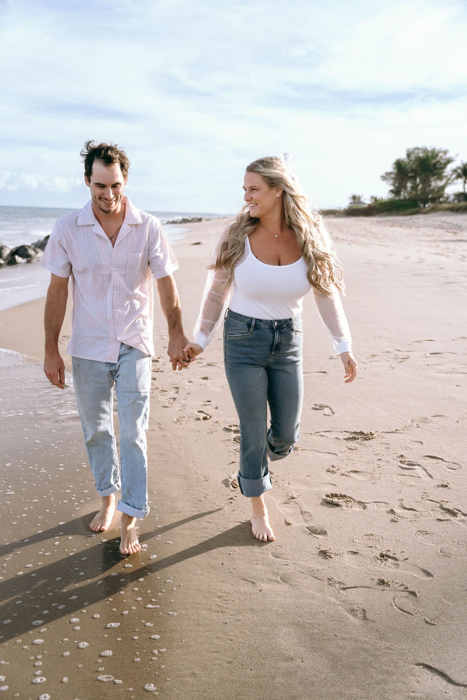 A couple walking barefoot along a sandy beach shore holding hands on a cloudy day.