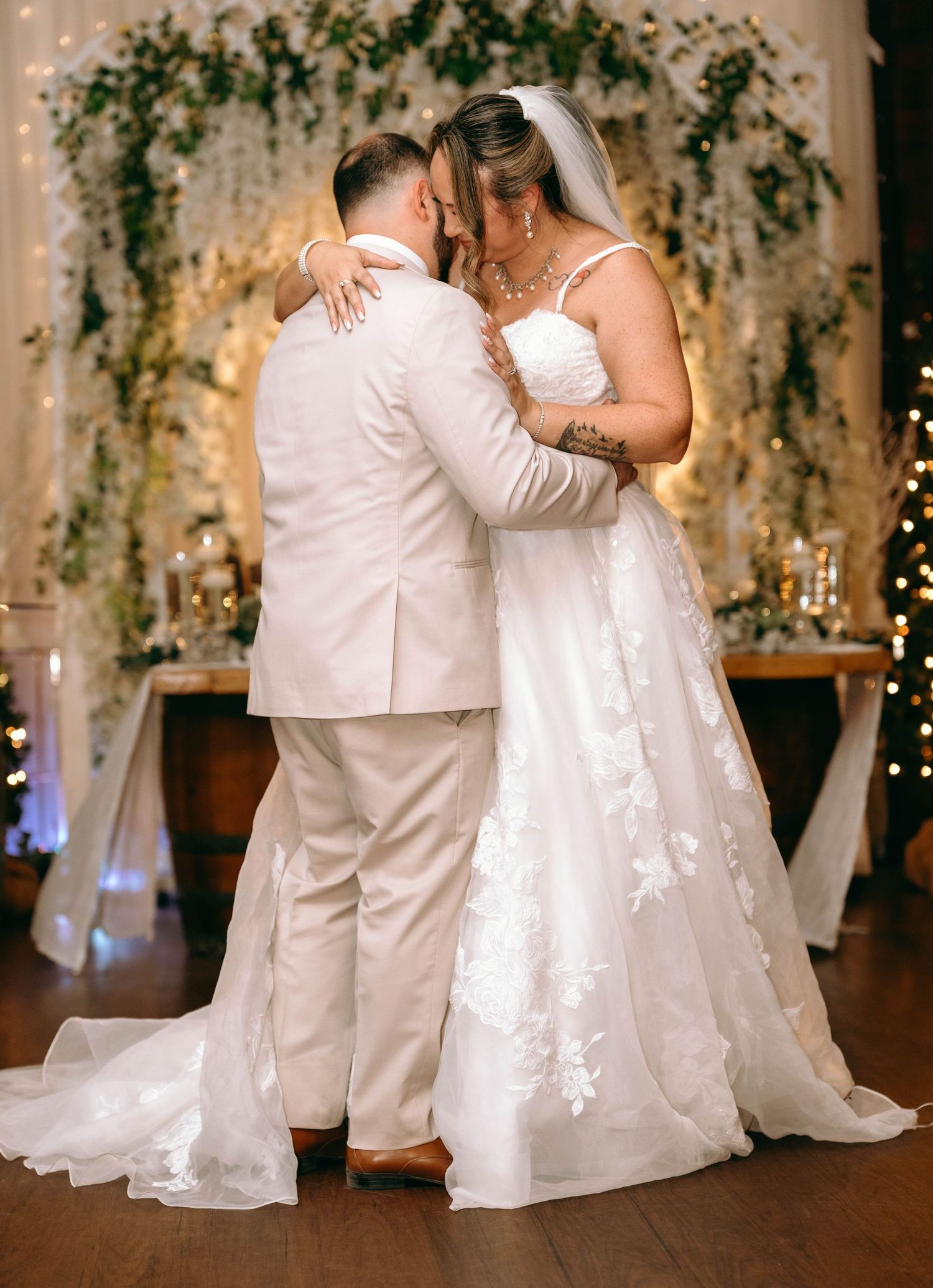 A couple shares their first dance at an elegant indoor wedding reception with floral arch decor and soft lighting.