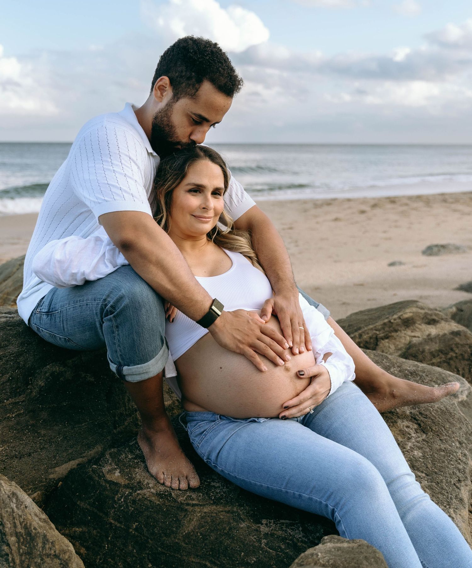 Romantic maternity photos on rocky beach with couple in casual denim and white attire.
