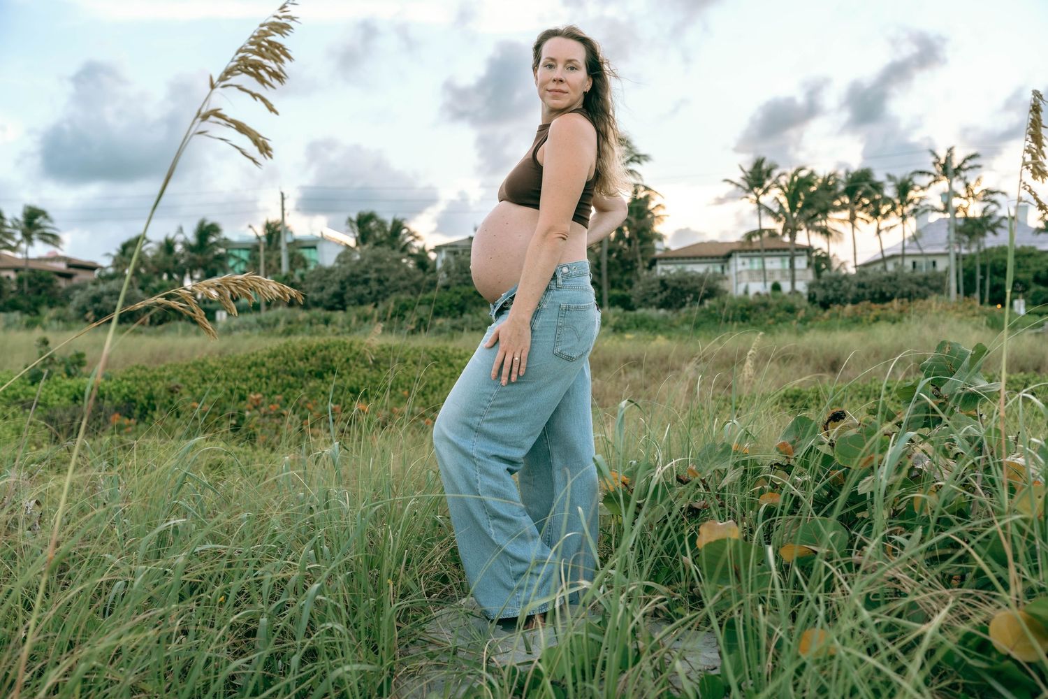 Pregnant woman in blue jeans and brown crop top poses outdoors in lush green field with tropical trees and cloudy sky.