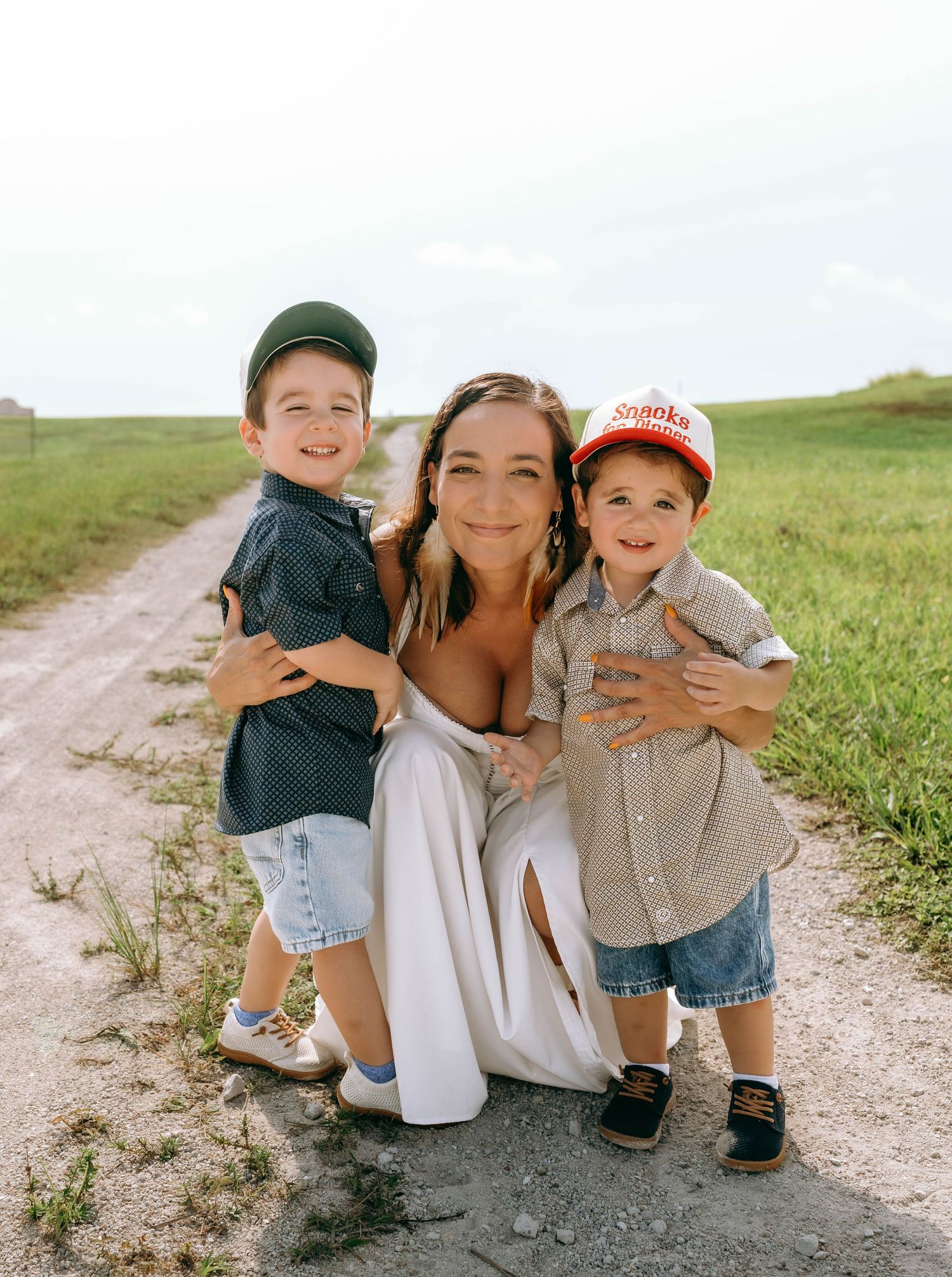 A woman crouches on a dirt path hugging two young boys in button-up shirts and caps, surrounded by green fields.