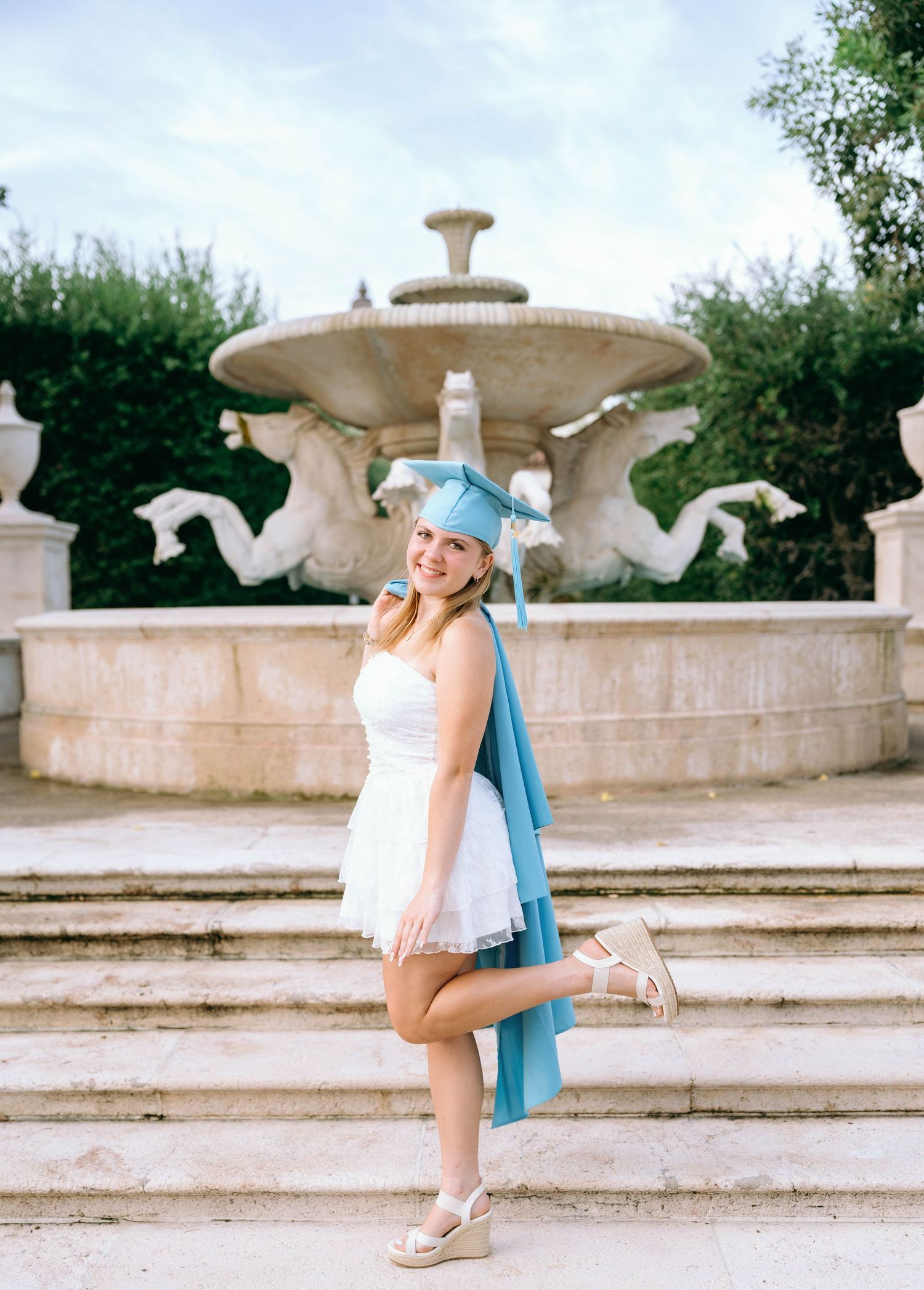 Smiling graduate in white dress and light blue cap poses by ornate stone fountain with sculptural details.